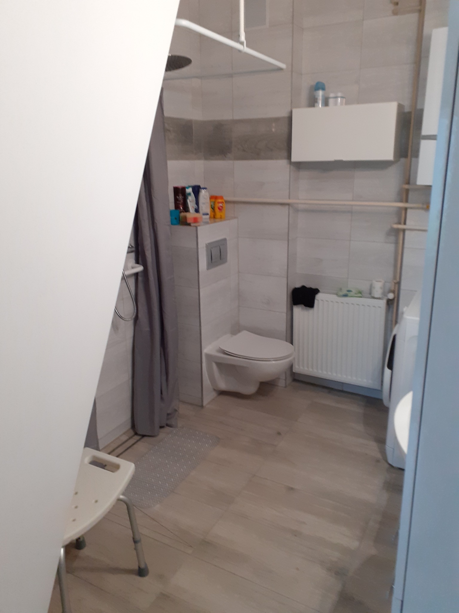 Widok na wnętrze łazienki z toaletą, grzejnikiem i pralką, częściowo zasłonięte drzwiami i zasłoną prysznicową.