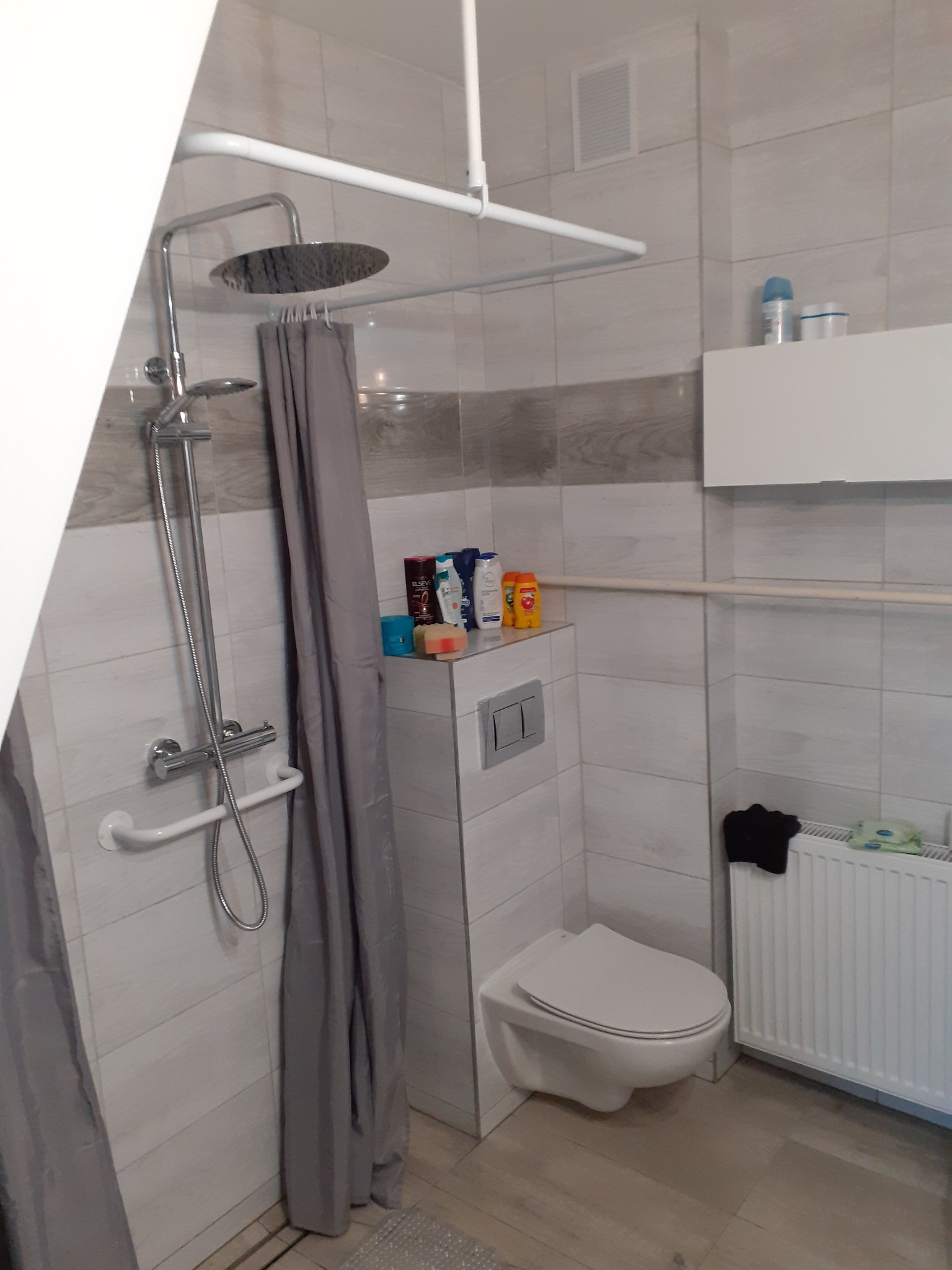 Wykończona łazienka w jasnych płytkach z kabiną prysznicową typu walk-in, deszczownicą, toaletą podwieszaną i grzejnikiem drabinkowym.