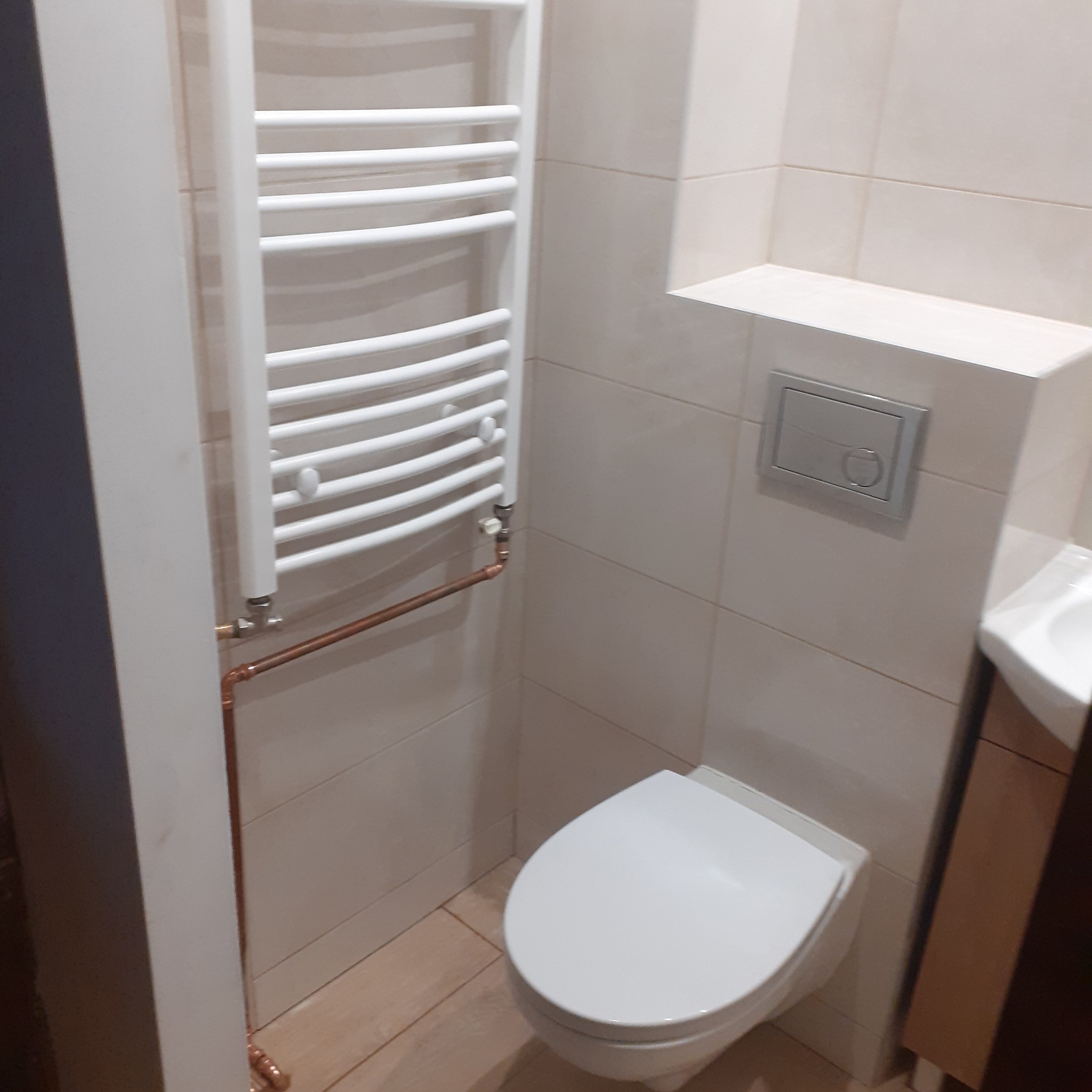 Nowoczesna łazienka z beżowymi płytkami, wiszącą toaletą, grzejnikiem drabinkowym i miedzianymi rurami instalacji grzewczej.