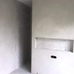 Artur Tudek wykończenia wnętrz usługi malarskie ,glazurnicze - Surowe ściany w szarym odcieniu z wnęką i gniazdkiem elektrycznym, widok na korytarz w tle, stan deweloperski.