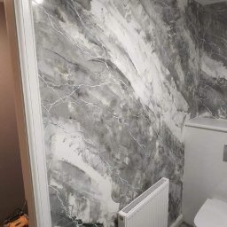 Artur Tudek wykończenia wnętrz usługi malarskie ,glazurnicze - Ściana w łazience z dekoracyjną fakturą w odcieniach szarości i bieli, przypominająca marmur z białymi żyłkami, widoczny grzejnik i toaleta w tle, podłoga wyłożona sześciokątnymi płytkami.