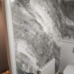 Ściana w łazience z dekoracyjną fakturą w odcieniach szarości i bieli, przypominająca marmur z białymi żyłkami, widoczny grzejnik i toaleta w tle, podłoga wyłożona sześciokątnymi płytkami.