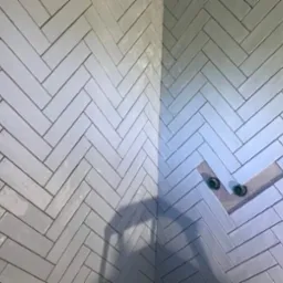 Ściana wyłożona błyszczącymi, szarymi płytkami ułożonymi w jodełkę, widoczne dwa zielone zawory wystające ze ściany w miejscu niedokończonego wykończenia.