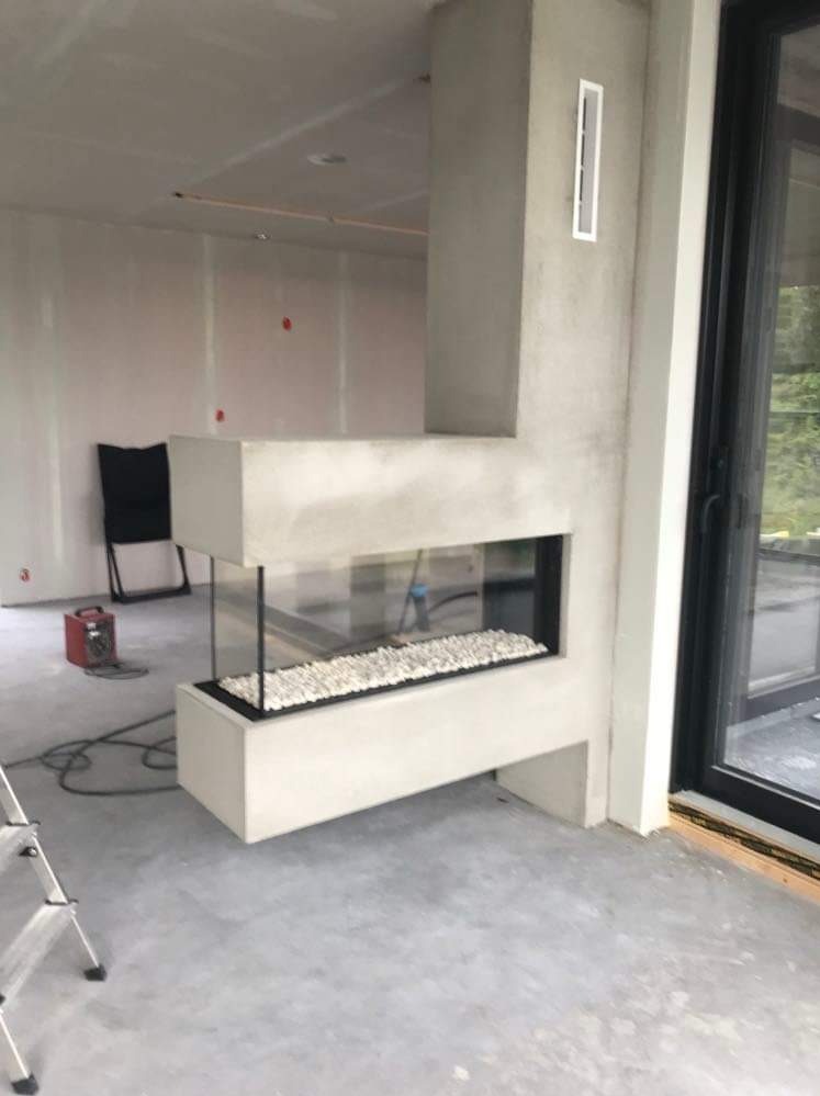 Wnętrze w trakcie wykończenia: minimalistyczny, betonowy kominek z białymi kamieniami dekoracyjnymi, widoczny grzejnik elektryczny i drabina malarska na szarym betonie.