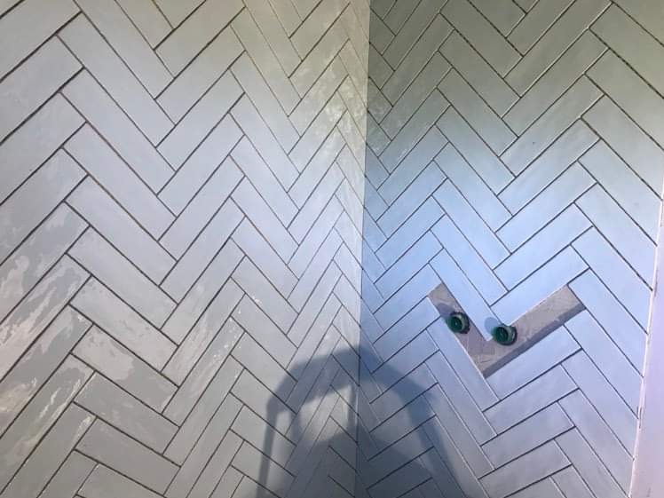 Ściana wyłożona jasnoszarymi, podłużnymi płytkami ułożonymi w jodełkę, z widocznymi dwoma zielonymi zaworami wystającymi ze ściany w miejscu niedokończonego układania płytek.