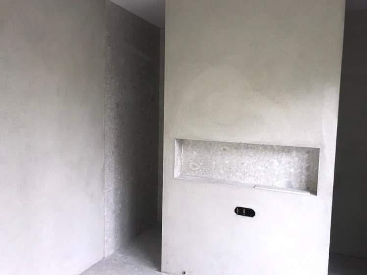 Surowe ściany w szarym odcieniu z wnęką i gniazdkiem elektrycznym, widok na korytarz w tle, stan deweloperski.