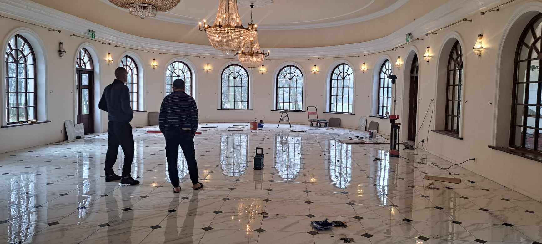 Przestronne, okrągłe wnętrze z marmurową podłogą w trakcie prac wykończeniowych; widoczne narzędzia, resztki zaprawy i dwóch mężczyzn w tle.