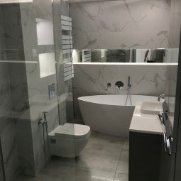 Nowoczesna łazienka z owalną wanną wolnostojącą, podwieszaną toaletą i umywalką nablatową na szarym blacie. Ściany wyłożone białymi płytkami imitującymi marmur z szarymi żyłami. Widoczny szklany...