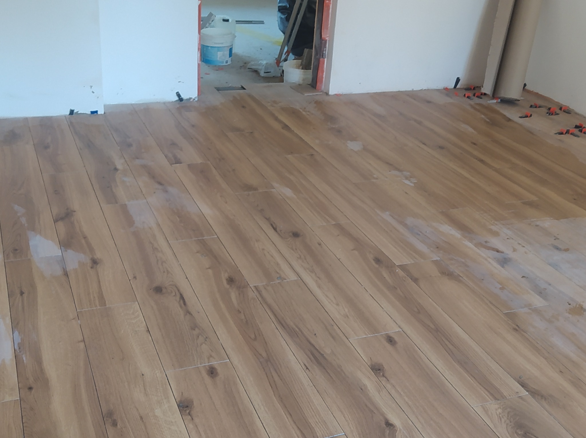 Układanie podłogi z płytek imitujących drewno, widoczne plastikowe kliny dystansowe, w tle wiadro i narzędzia budowlane.