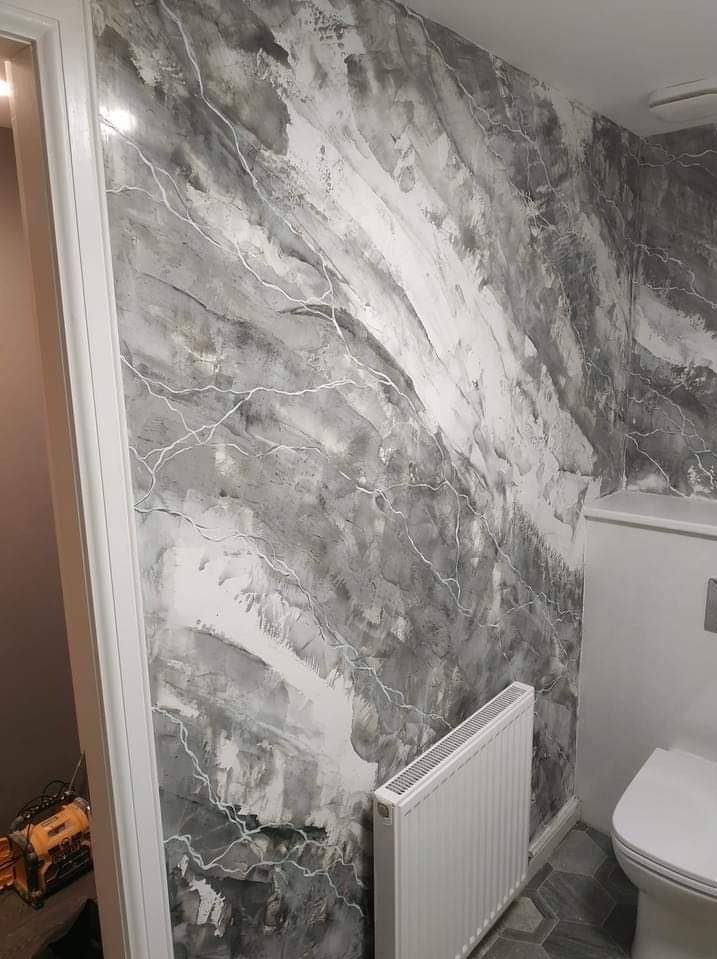 Ściana w łazience z dekoracyjną fakturą w odcieniach szarości i bieli, przypominająca marmur z białymi żyłkami, widoczny grzejnik i toaleta w tle, podłoga wyłożona sześciokątnymi płytkami.