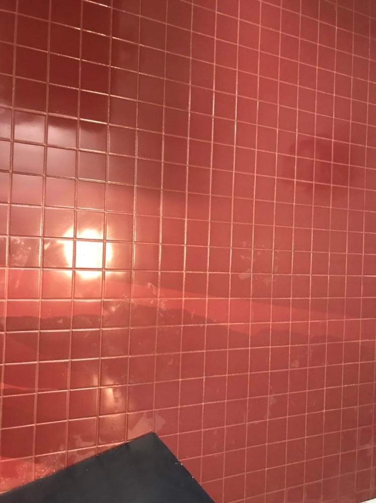 Ściana wyłożona błyszczącymi, kwadratowymi, czerwonymi płytkami ceramicznymi; widoczne odbicie światła i fragment czarnego narzędzia w dolnym rogu.