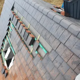 Układanie dachówki ceramicznej w dwóch odcieniach szarości i brązu, widoczne łaty drewniane i membrana dachowa, fragment dachu w trakcie prac.