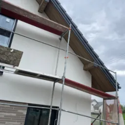 Dom w trakcie prac dekarskich: widoczne rusztowanie, fragment dachu z grafitową dachówką i drewnianą podbitką. Ściana budynku częściowo otynkowana, z oknami.