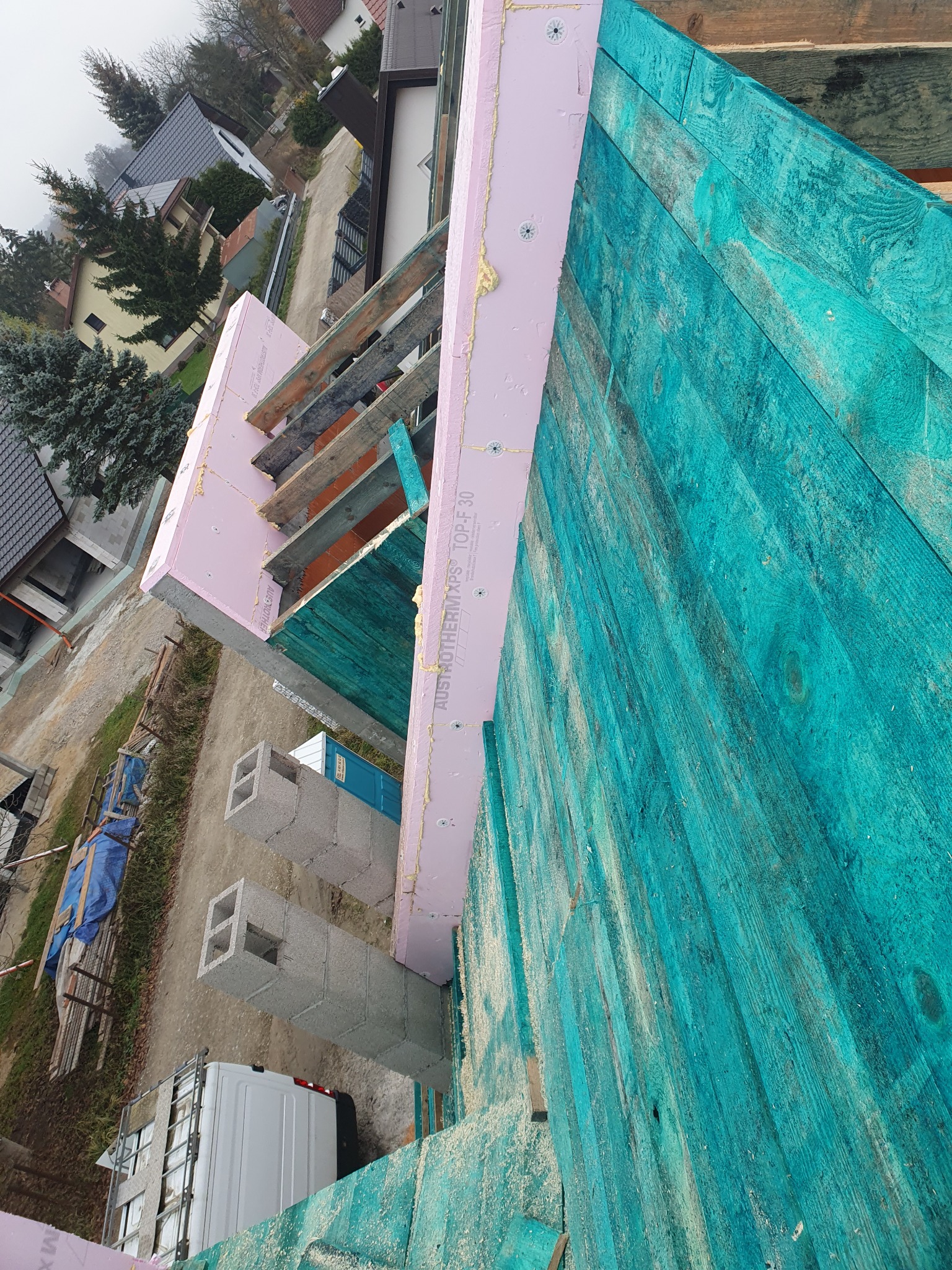 Widok z góry na fragment dachu w trakcie budowy: turkusowe deski, różowa izolacja termiczna Austrotherm, drewniana więźba, w tle zabudowa jednorodzinna i samochód dostawczy.