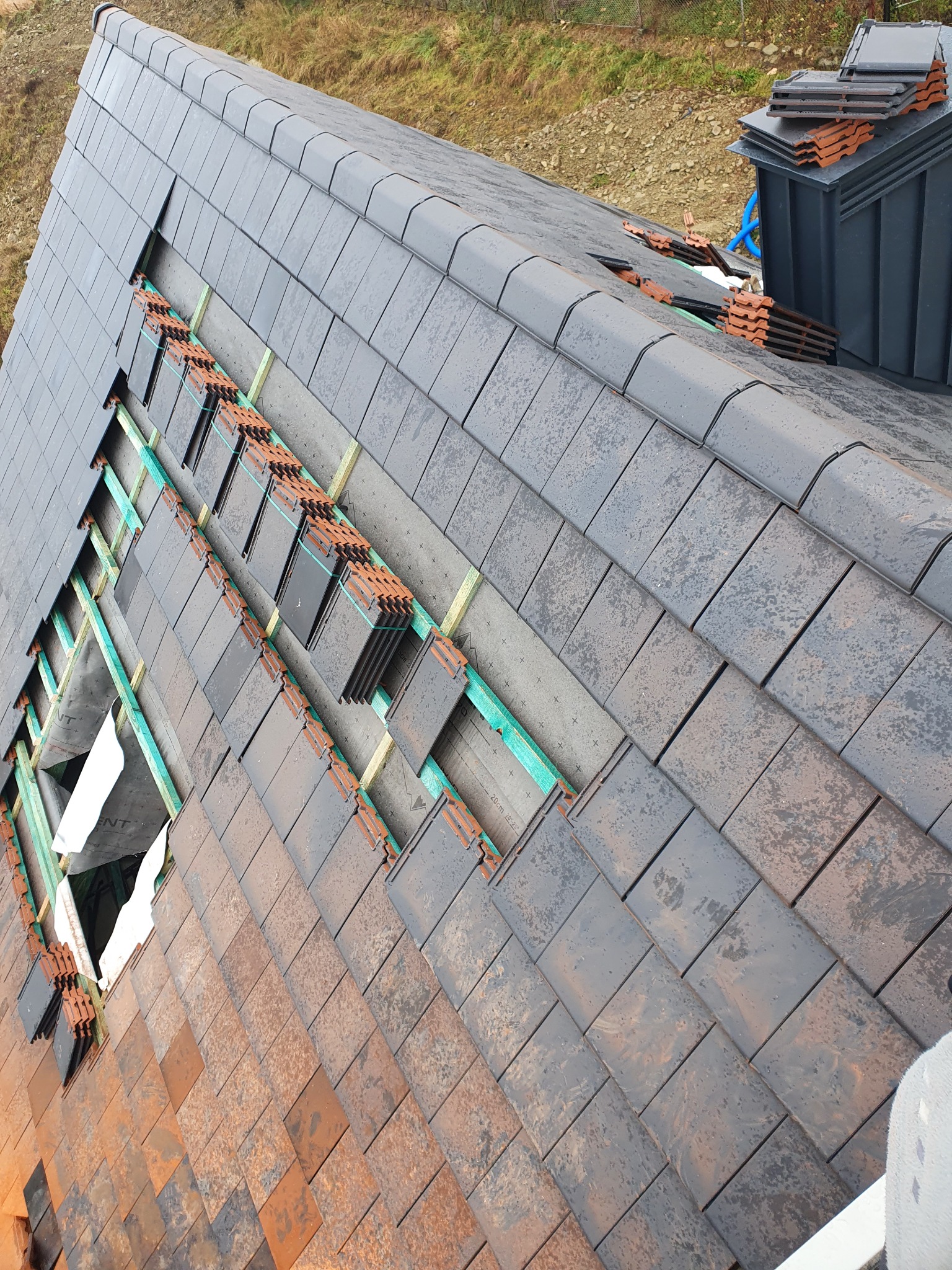 Układanie dachówki ceramicznej w dwóch odcieniach szarości i brązu, widoczne łaty drewniane i membrana dachowa, fragment dachu w trakcie prac.