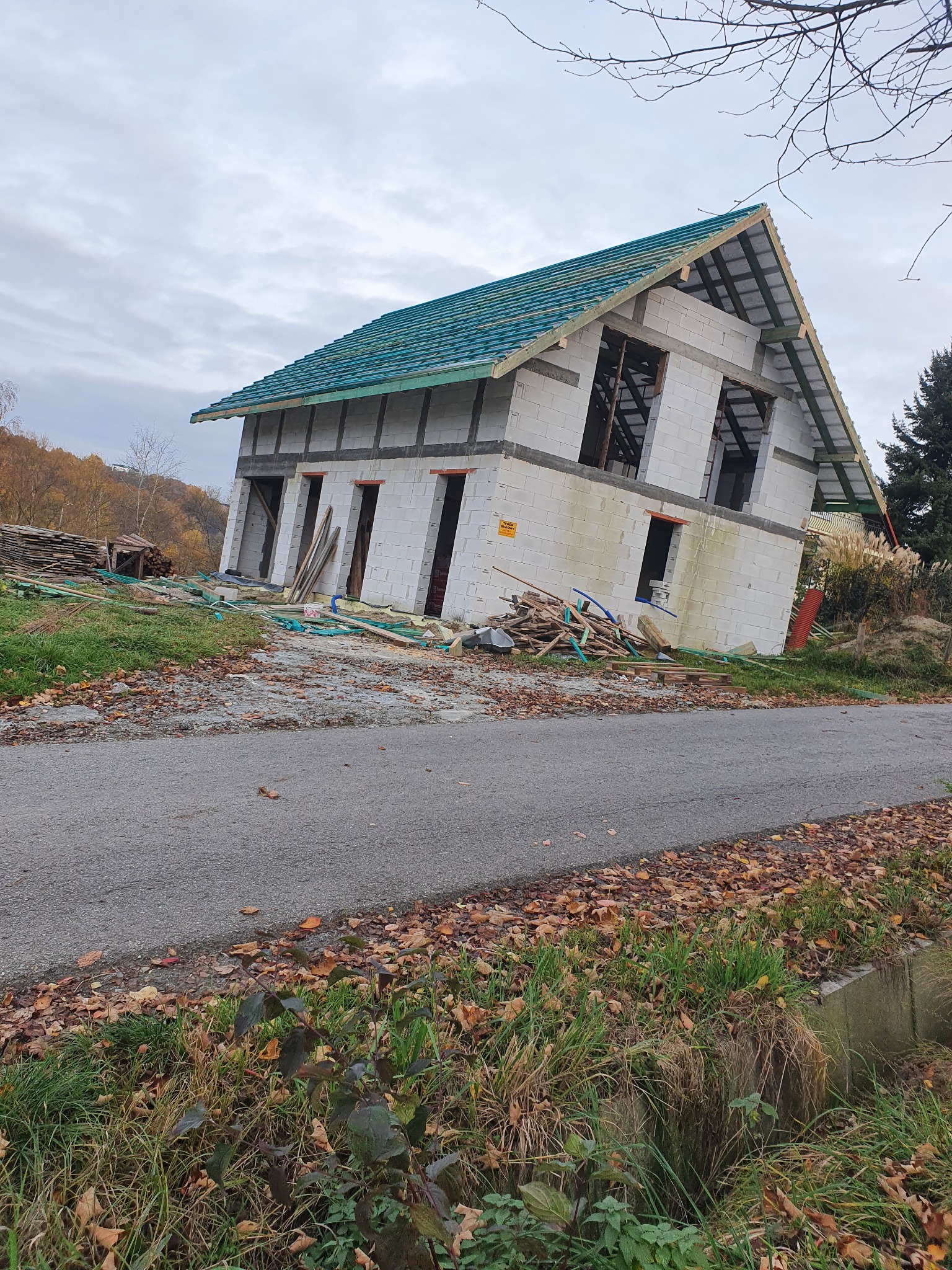 Niedokończony budynek z zielonym dachem w trakcie budowy, widoczne pustaki, więźba dachowa i sterta materiałów budowlanych obok drogi.
