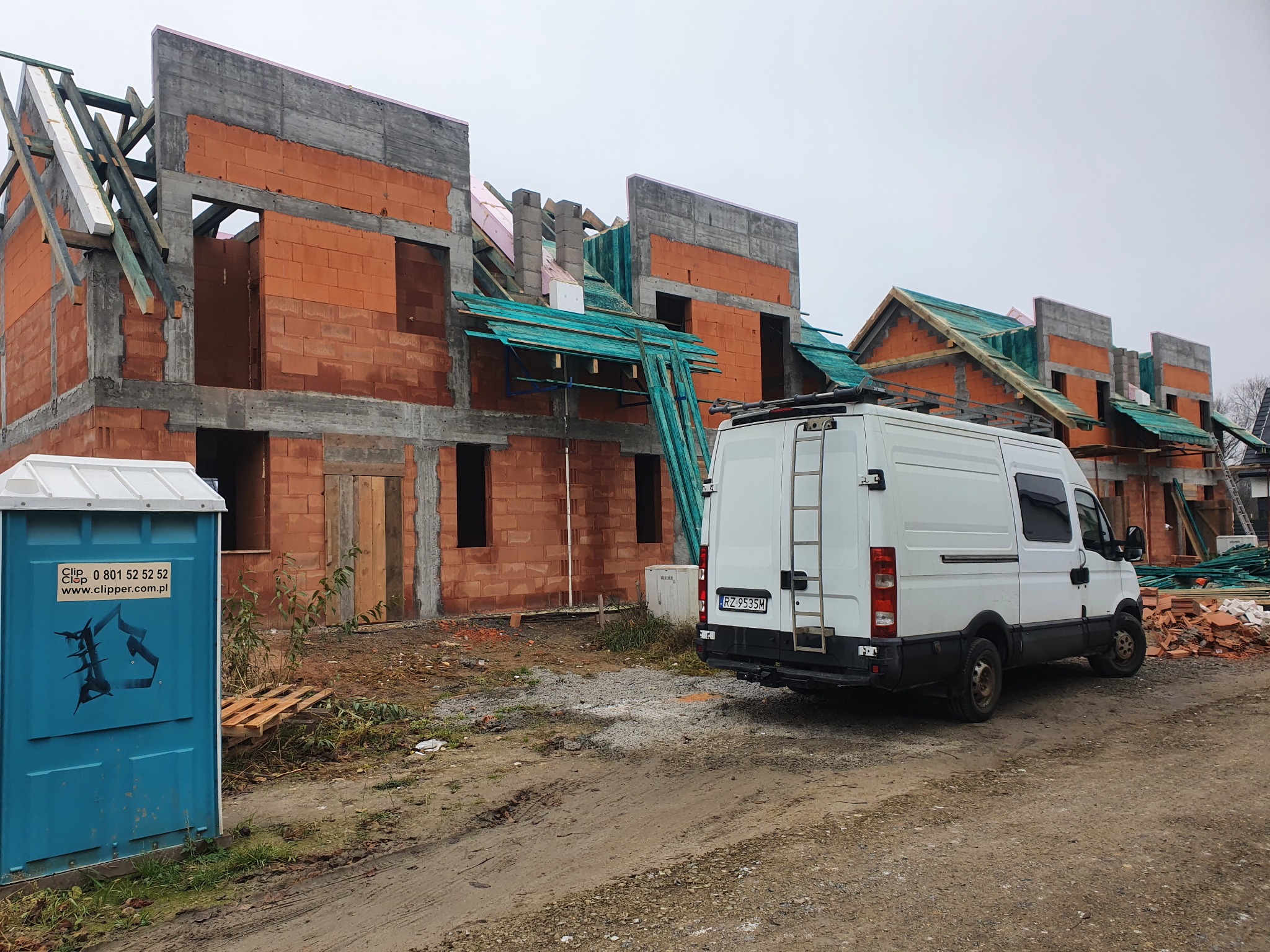 Budowa domów w stanie surowym otwartym z widoczną konstrukcją dachu, na pierwszym planie toaleta przenośna i samochód dostawczy.