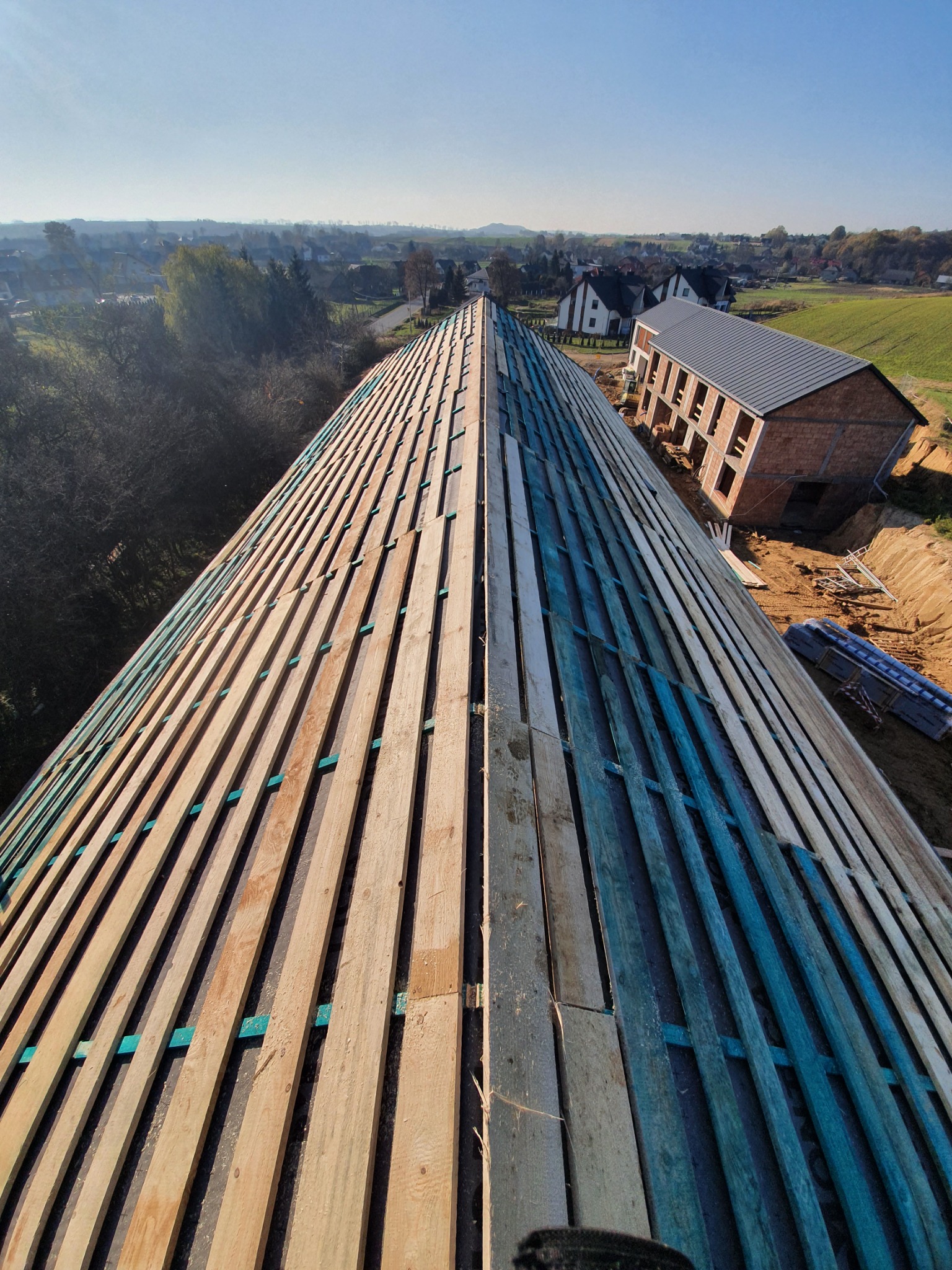 Wysoki kąt widzenia na konstrukcję dachu z drewnianymi łatami w dwóch odcieniach, z widocznym budynkiem w stanie surowym otwartym i rozległym krajobrazem w tle.