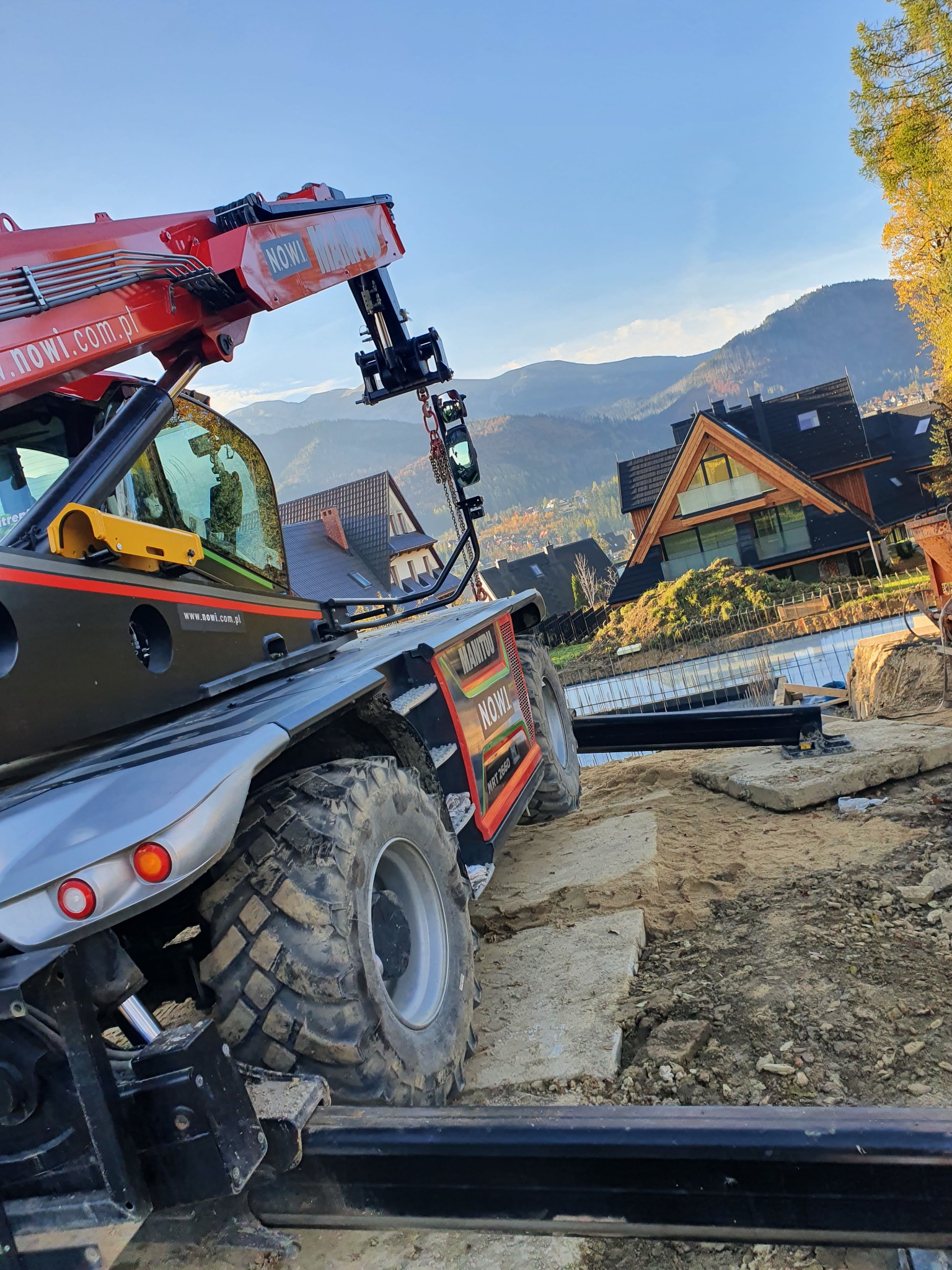 Czerwono-szary podnośnik teleskopowy Manitou na placu budowy z widokiem na góry i dom w stylu zakopiańskim w tle, w słoneczny dzień.