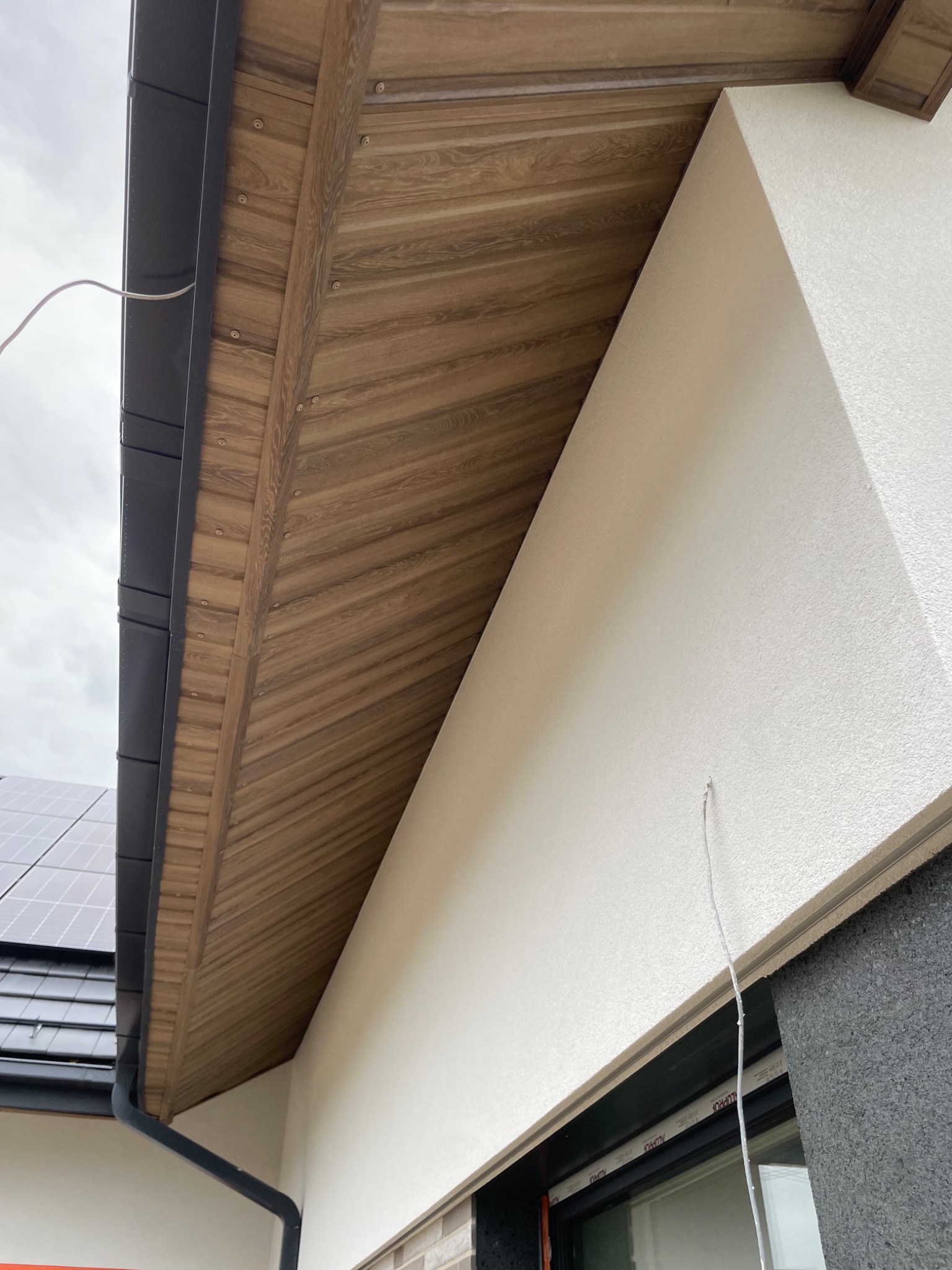 Detale wykończenia okapu z drewnianą podbitką i czarną rynną, widoczne panele słoneczne na dachu w tle. Nowoczesne wykończenie elewacji budynku.