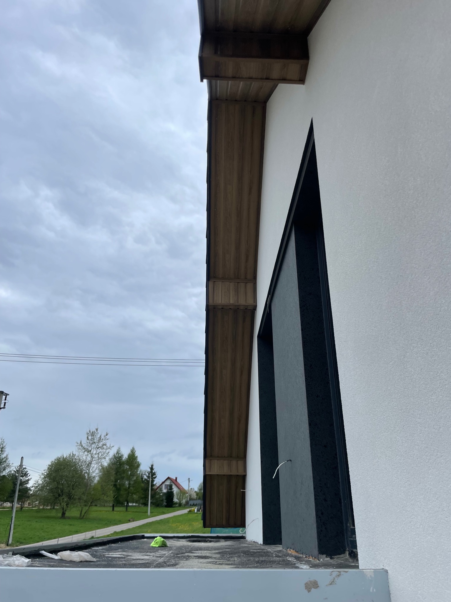 Fragment elewacji budynku z drewnianym okapem i czarną ramą okienną, widok z dołu na tle pochmurnego nieba i zieleni. Detal wykończenia dachu i ściany.