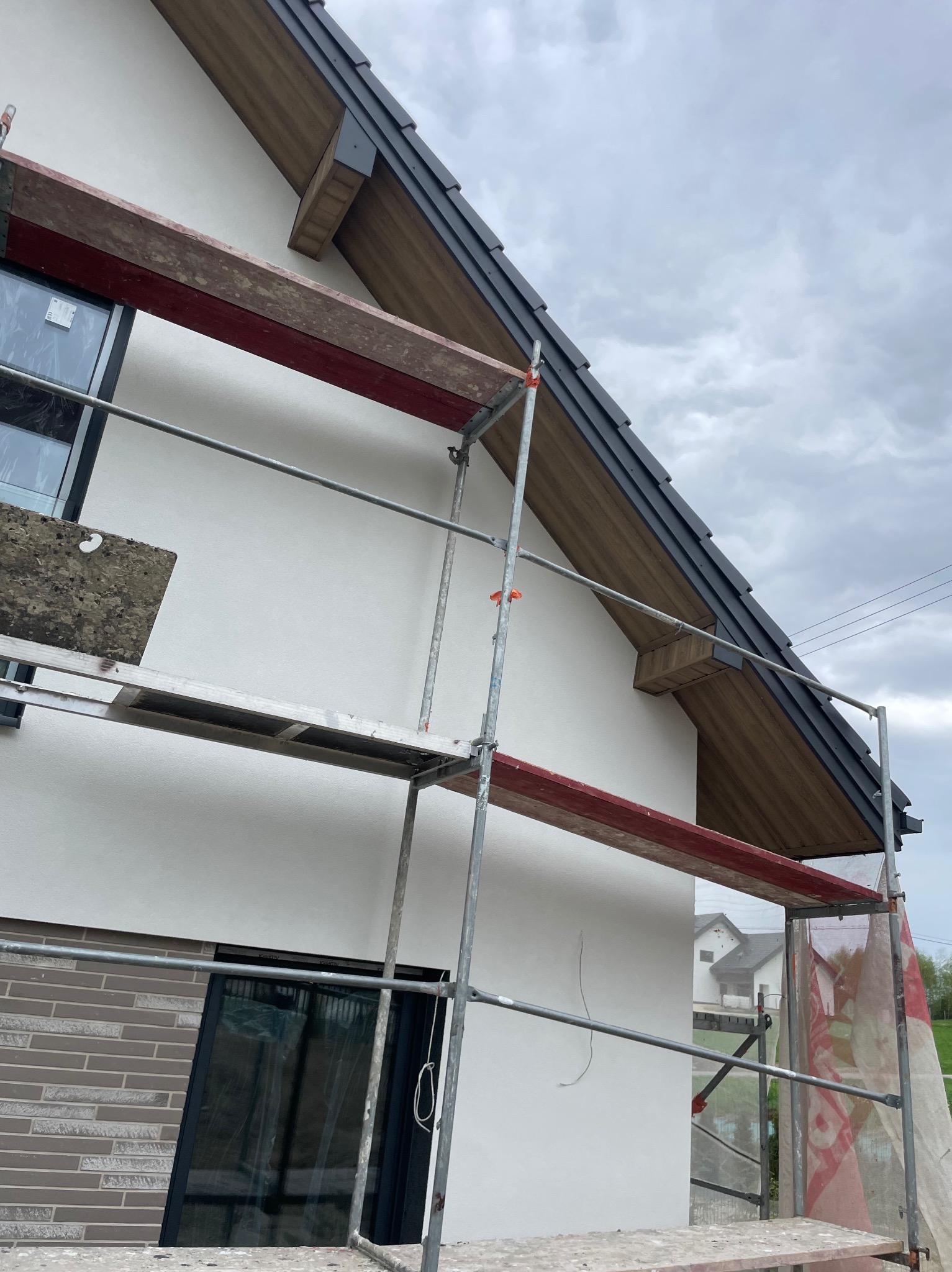 Dom w trakcie prac dekarskich: widoczne rusztowanie, fragment dachu z grafitową dachówką i drewnianą podbitką. Ściana budynku częściowo otynkowana, z oknami.