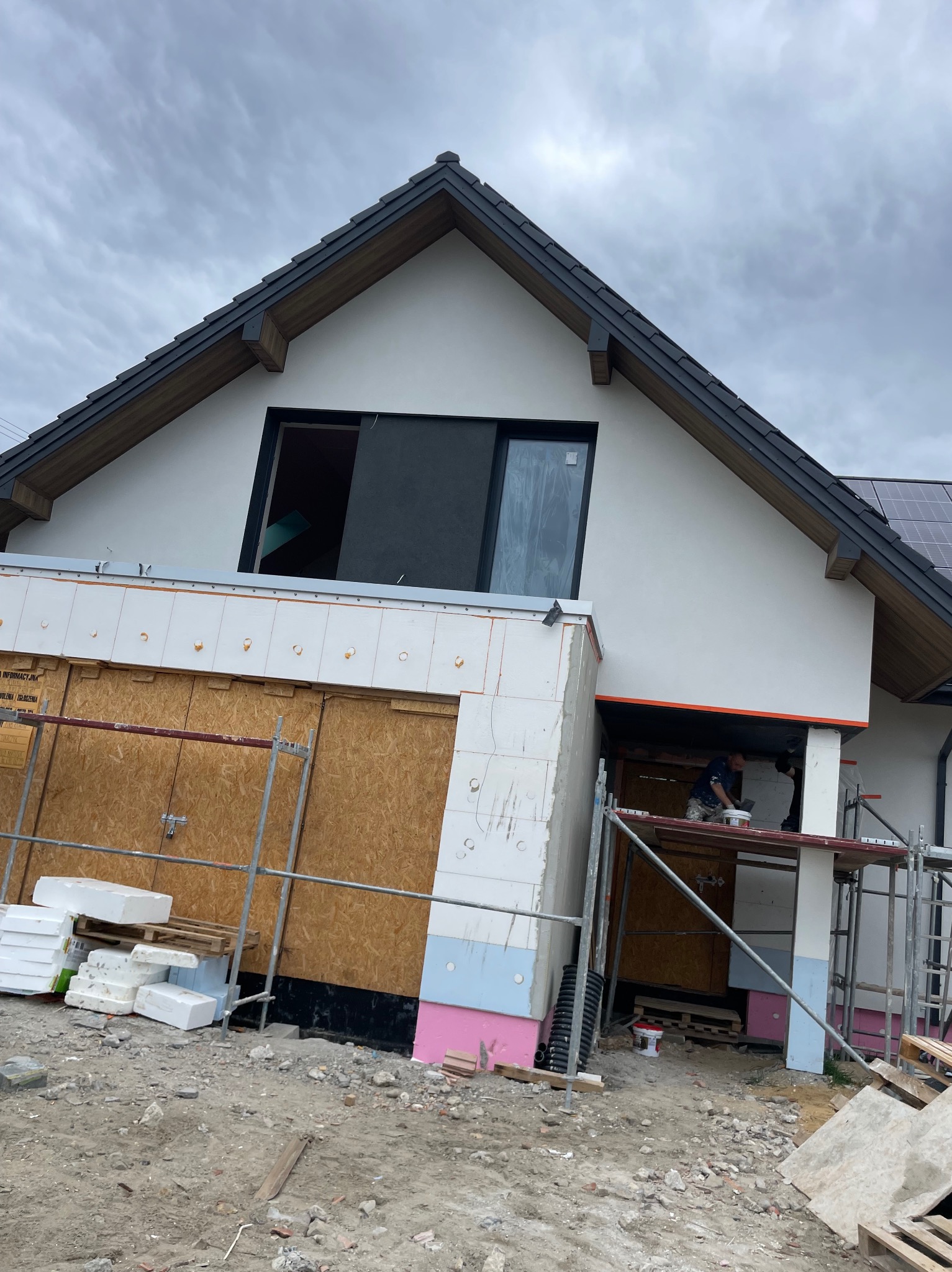 Dom w trakcie prac elewacyjnych: ocieplanie i tynkowanie. Widoczne rusztowania, styropian i pracownicy przy pracy. Dach z ciemnej dachówki.