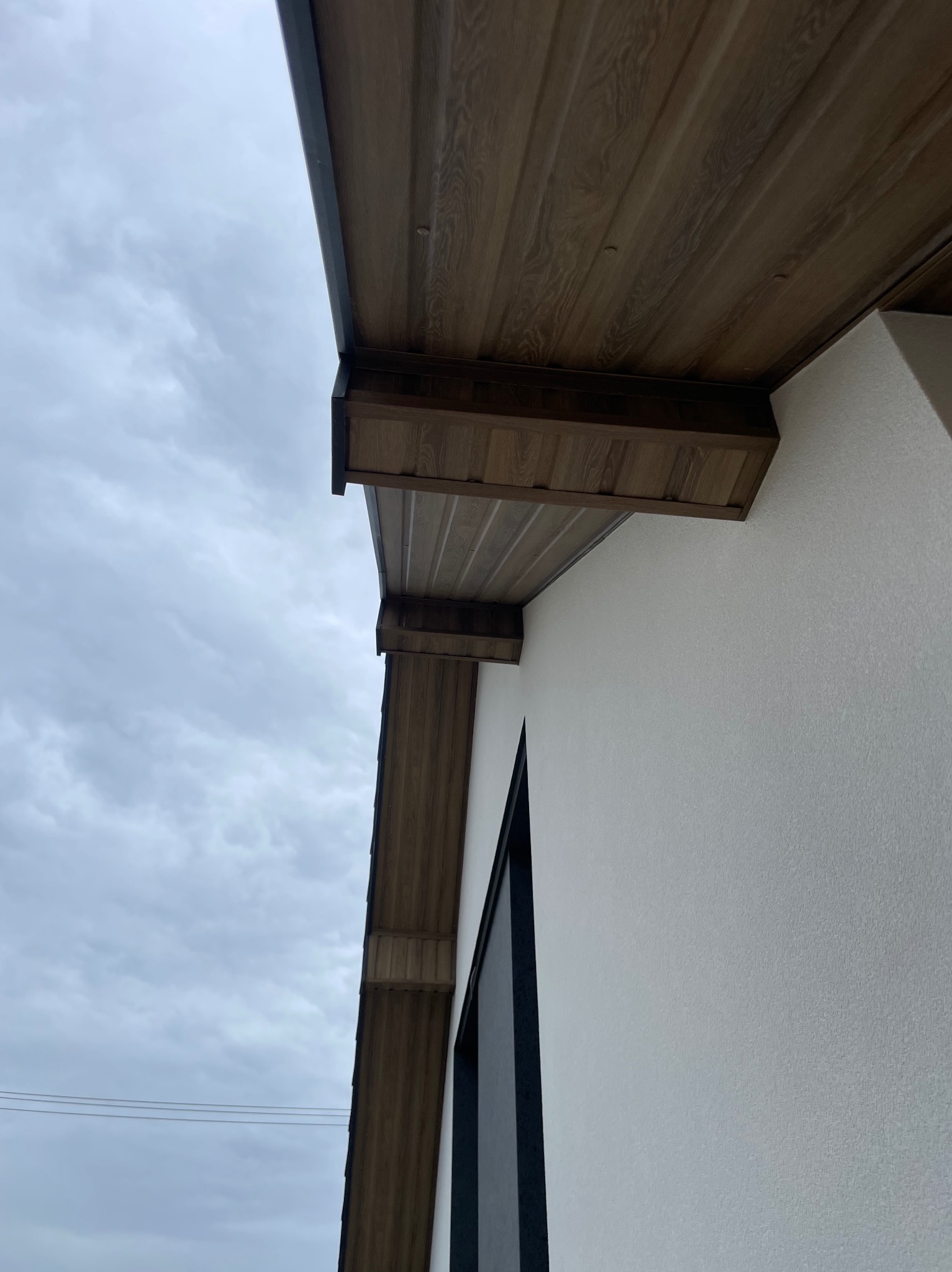 Drewniana podbitka dachowa z widoczną strukturą drewna, zamontowana przy białej ścianie budynku na tle pochmurnego nieba. Estetyczne wykończenie dachu.