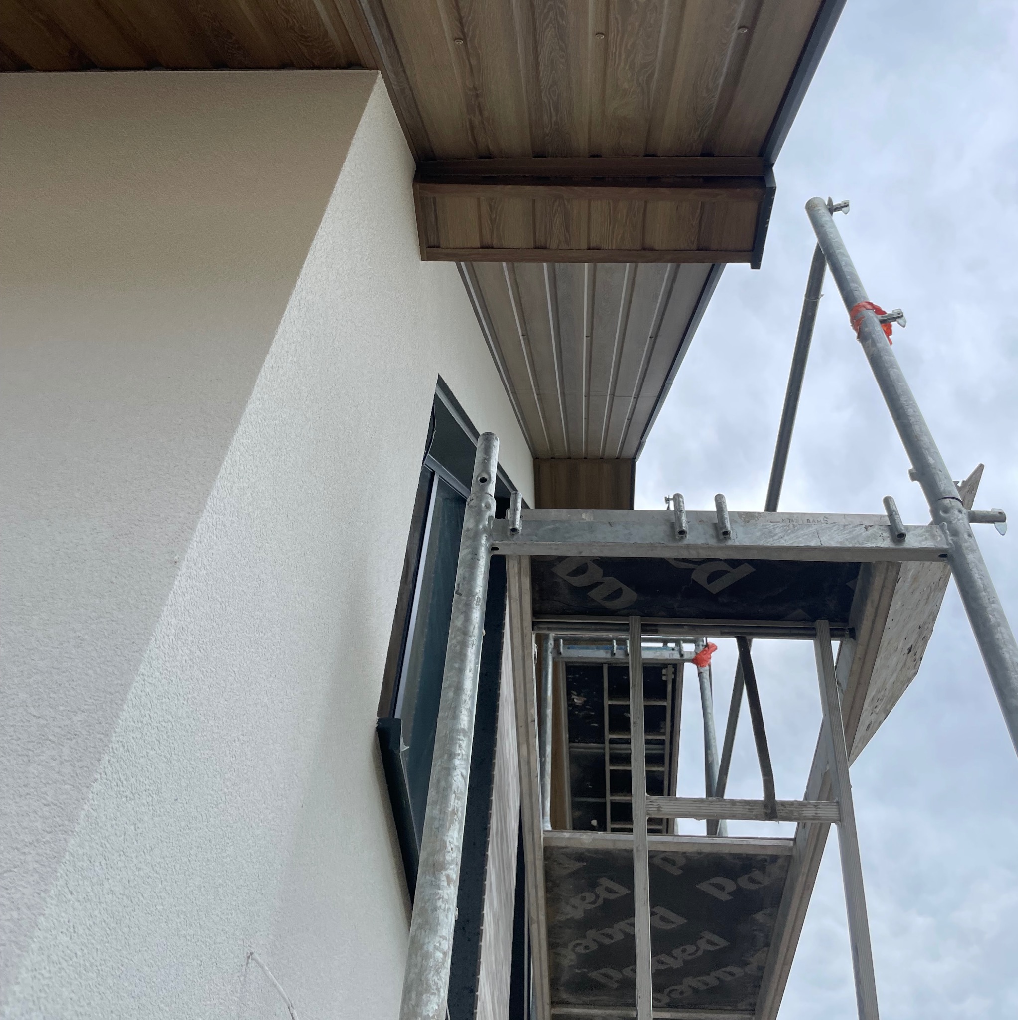 Montaż podbitki dachowej z rusztowania przy ścianie budynku. Widoczne panele w kolorze drewna i fragment okna. Pochmurne niebo w tle.