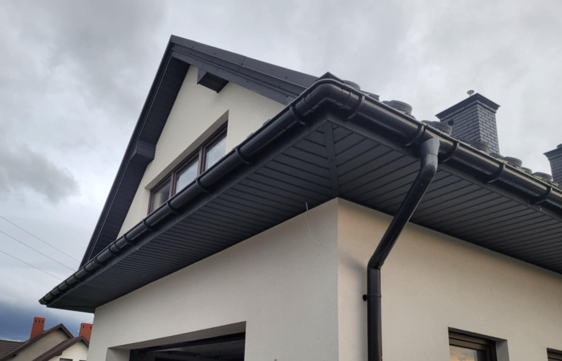 Fragment elewacji domu z zamontowanymi, czarnymi rynnami i podbitką dachową. Widoczny komin z szarej cegły. Pochmurne niebo w tle.