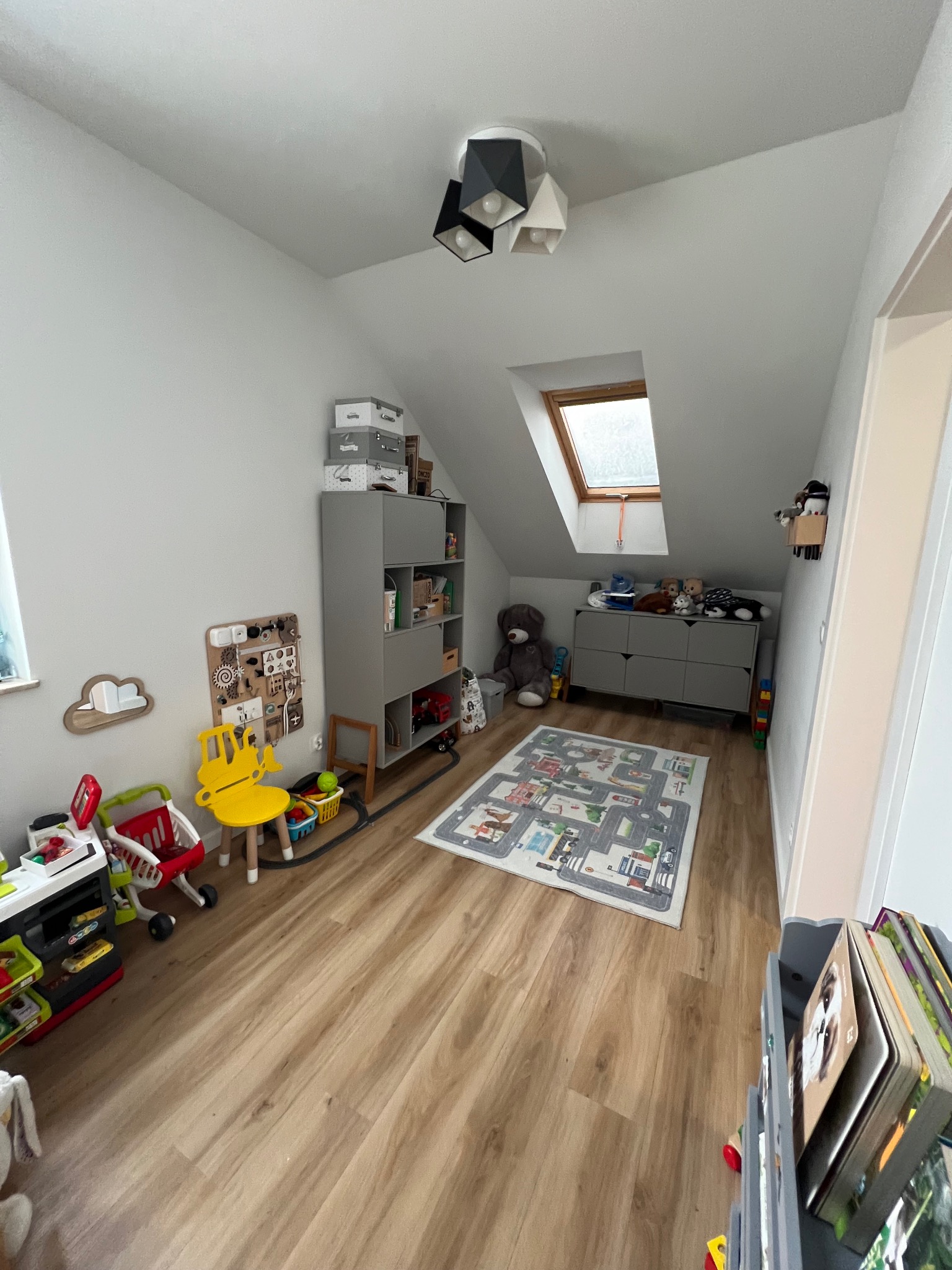 Uporządkowany pokój dziecięcy na poddaszu z drewnianą podłogą, szarymi meblami, kolorowymi zabawkami i dywanem z motywem ulic, oświetlony naturalnym światłem z okna dachowego.
