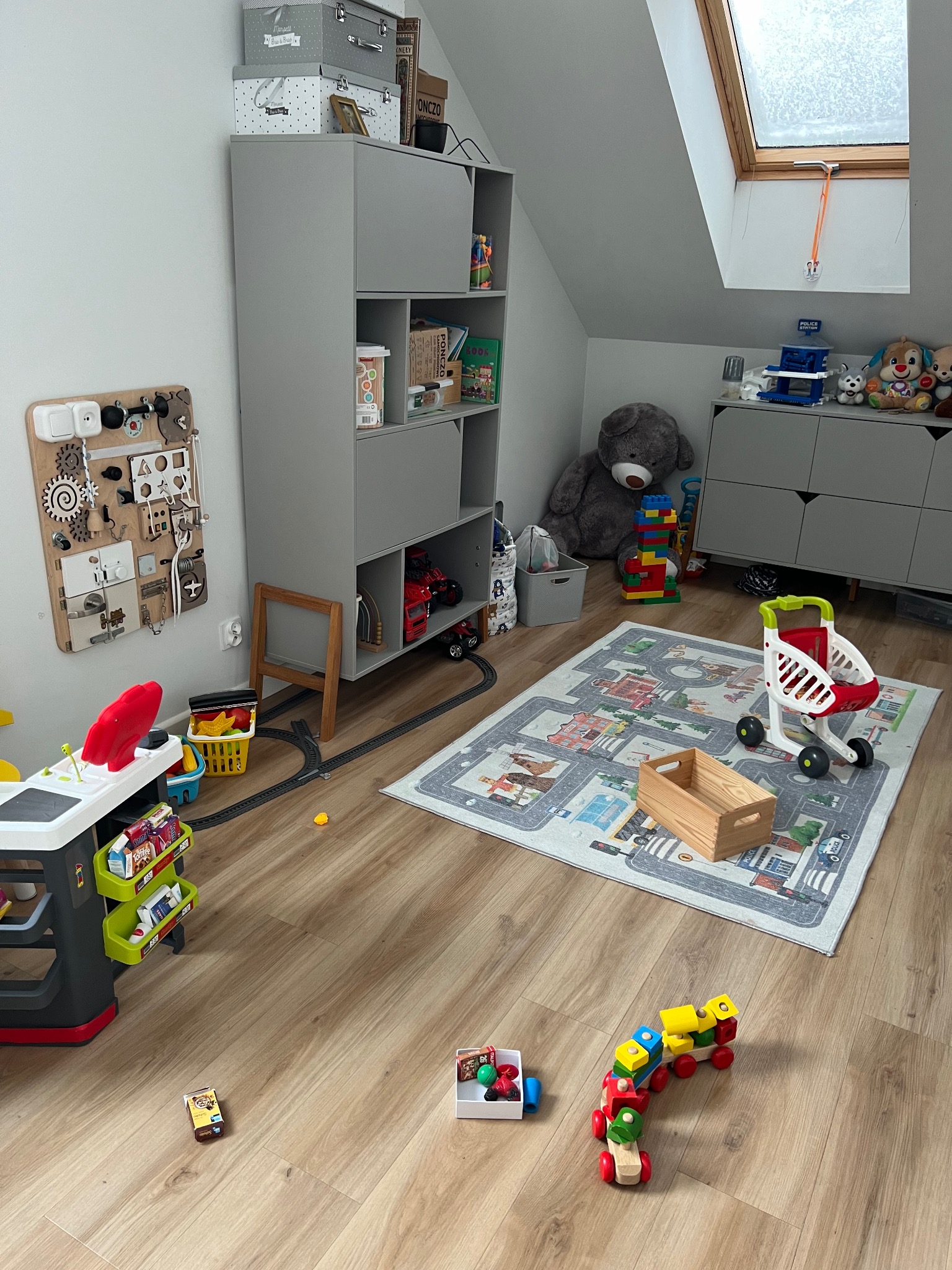 Pokój dziecięcy na poddaszu z drewnianą podłogą, szarymi meblami, dywanem z motywem ulic, zabawkami, w tym drewnianym pociągiem, wózkiem sklepowym i interaktywną tablicą na ścianie.