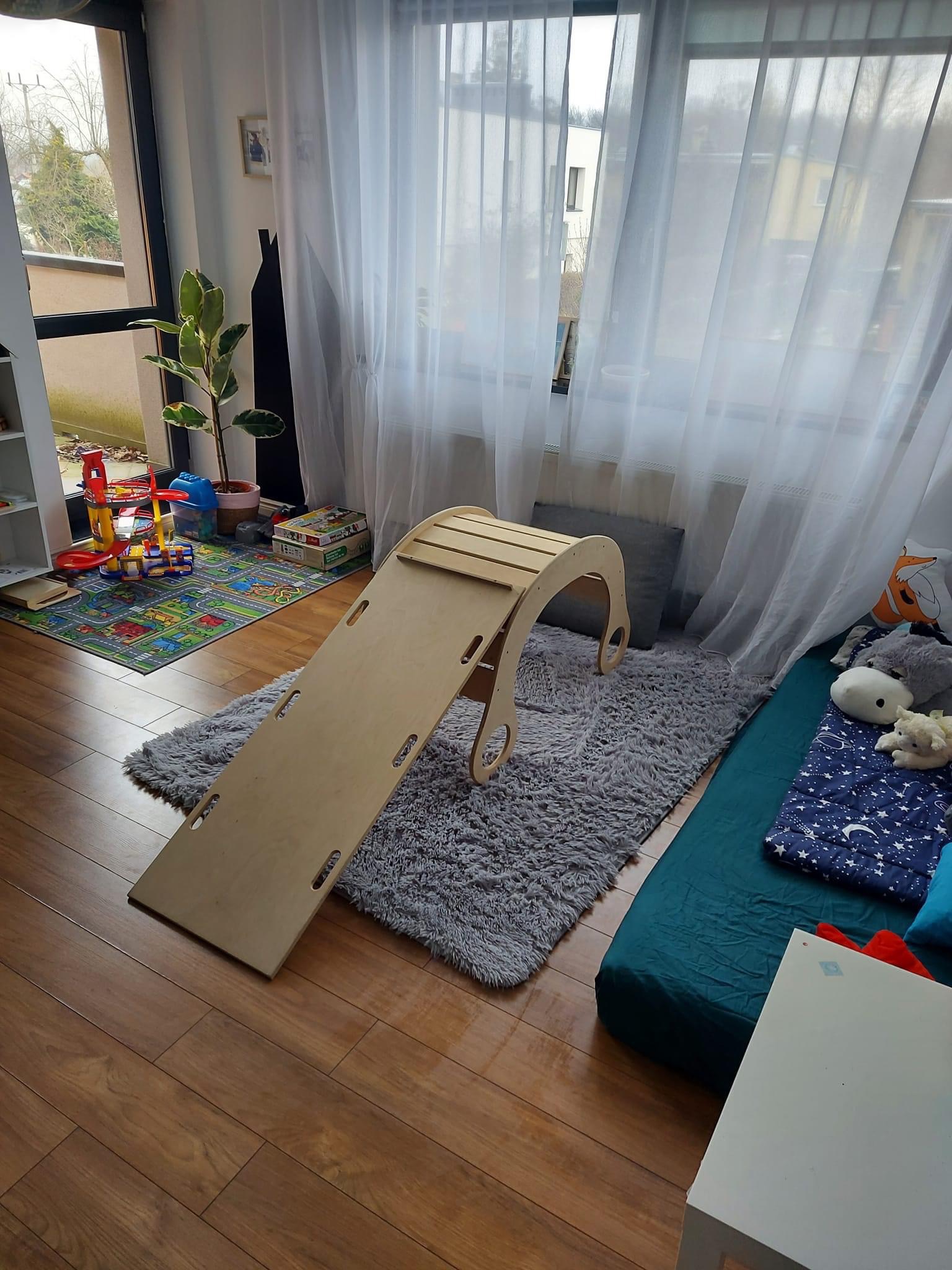 Drewniana zjeżdżalnia dla dzieci ustawiona na puszystym szarym dywanie w pokoju zabaw, obok widoczny materac w kolorze morskim z poduszkami i wzorem w gwiazdki, w tle białe zasłony i zabawki.