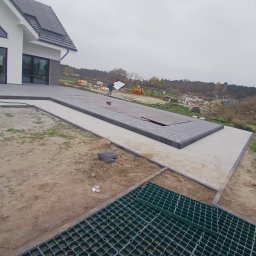 Przemyslaw Szota - Montaż nawierzchni z kostki brukowej w nowoczesnym ogrodzie, widoczne elementy drenażu w postaci zielonej kratki, w tle pracownik z płytą izolacyjną.