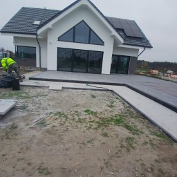 Przemyslaw Szota - Nowoczesny dom jednorodzinny z szarą kostką brukową na podjeździe i ścieżce, pracownik w odblaskowej kamizelce układa kolejne elementy, widoczne narzędzia i materiały budowlane.