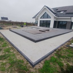 Przemyslaw Szota - Nowoczesny taras z szarej kostki brukowej obramowany ciemnoszarym obrzeżem, częściowo przykrywający basen, na tle białego domu z dużymi oknami i szarym dachem z panelami słonecznymi, widoczny...