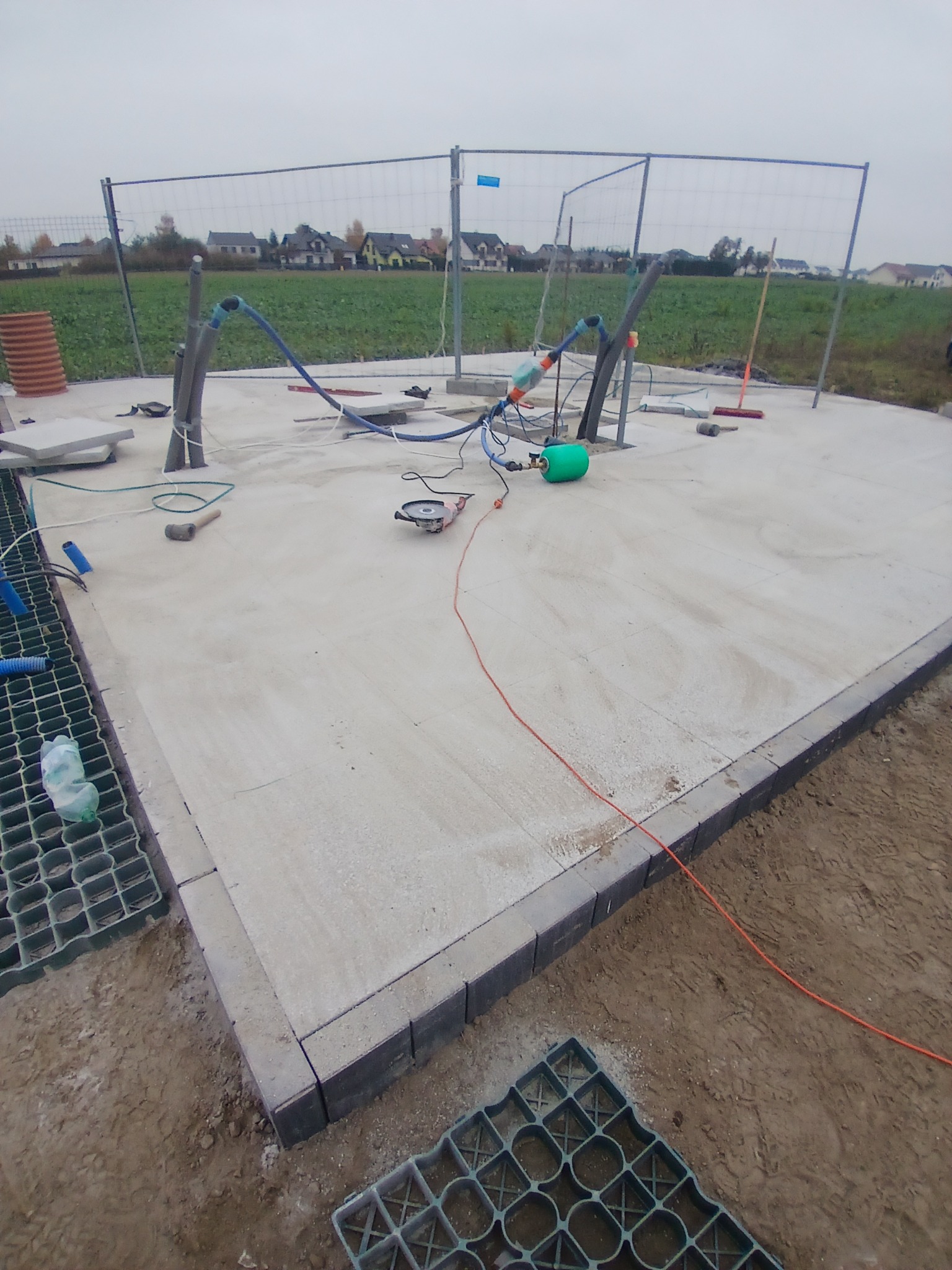 Przygotowany betonowy fundament z obrzeżem z kostki, widoczne rury instalacyjne, narzędzia i elementy drenażowe, ogrodzenie tymczasowe w tle.