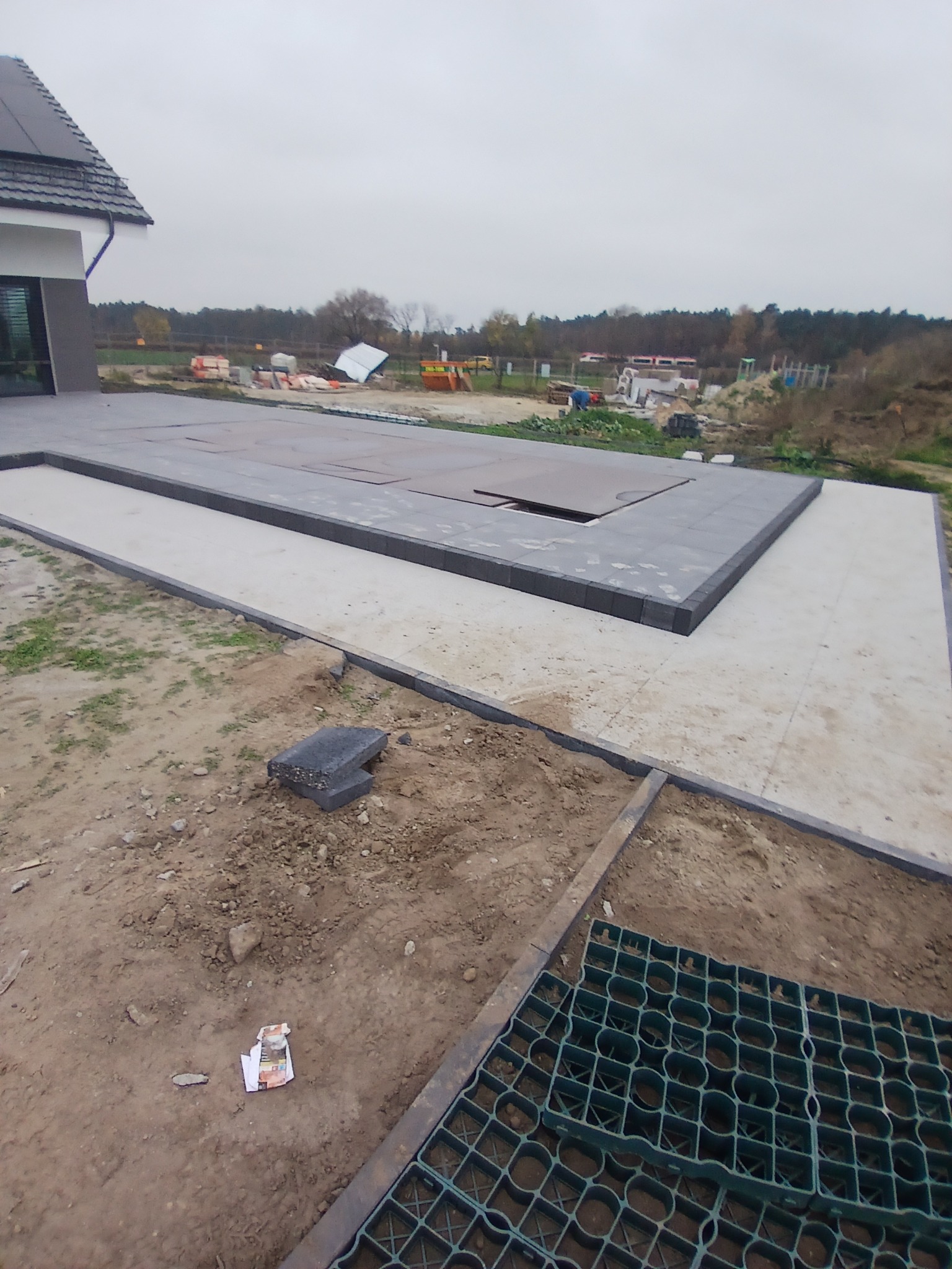 Betonowa platforma z obramowaniem z ciemnej kostki w trakcie budowy, widoczna zielona geokrata i zaplecze budowy w tle, pochmurny dzień.