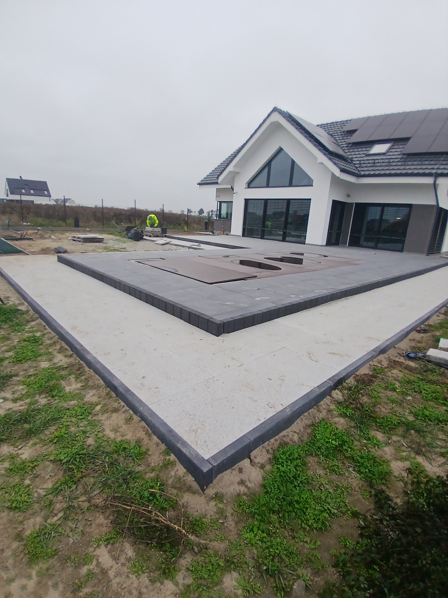 Nowoczesny taras z szarej kostki brukowej obramowany ciemnoszarym obrzeżem, częściowo przykrywający basen, na tle białego domu z dużymi oknami i szarym dachem z panelami słonecznymi, widoczny...