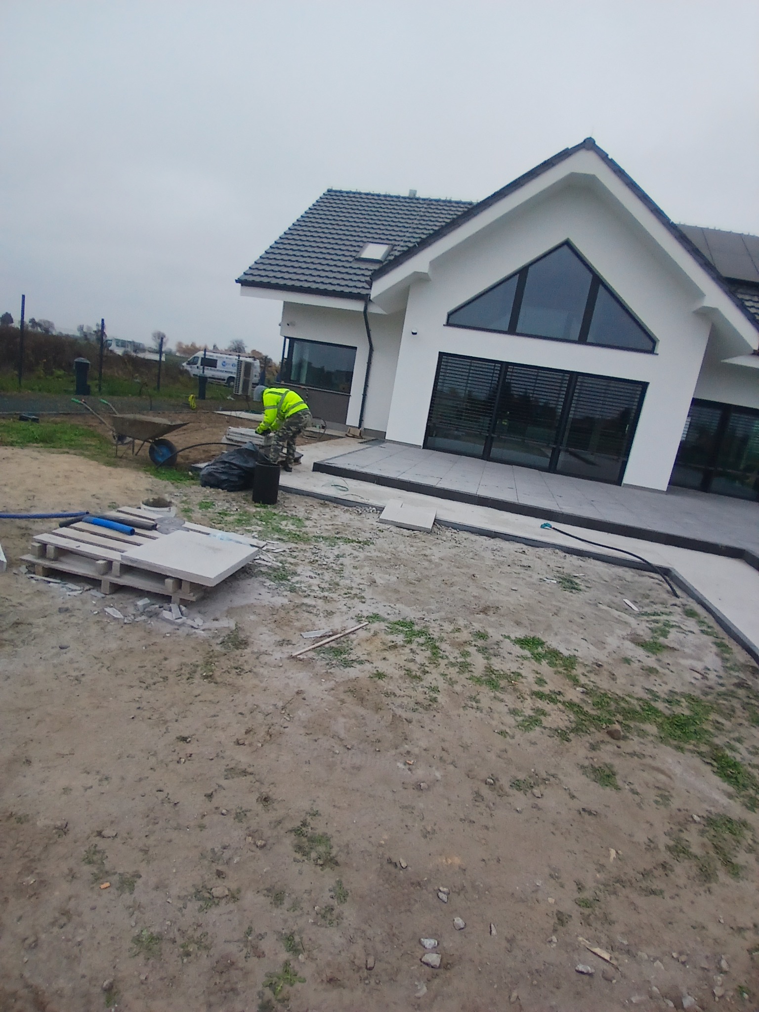 Pracownik w odblaskowej kamizelce przygotowuje teren przed nowoczesnym domem z szarymi płytami tarasowymi, widoczne palety z płytami, taczka i węże.