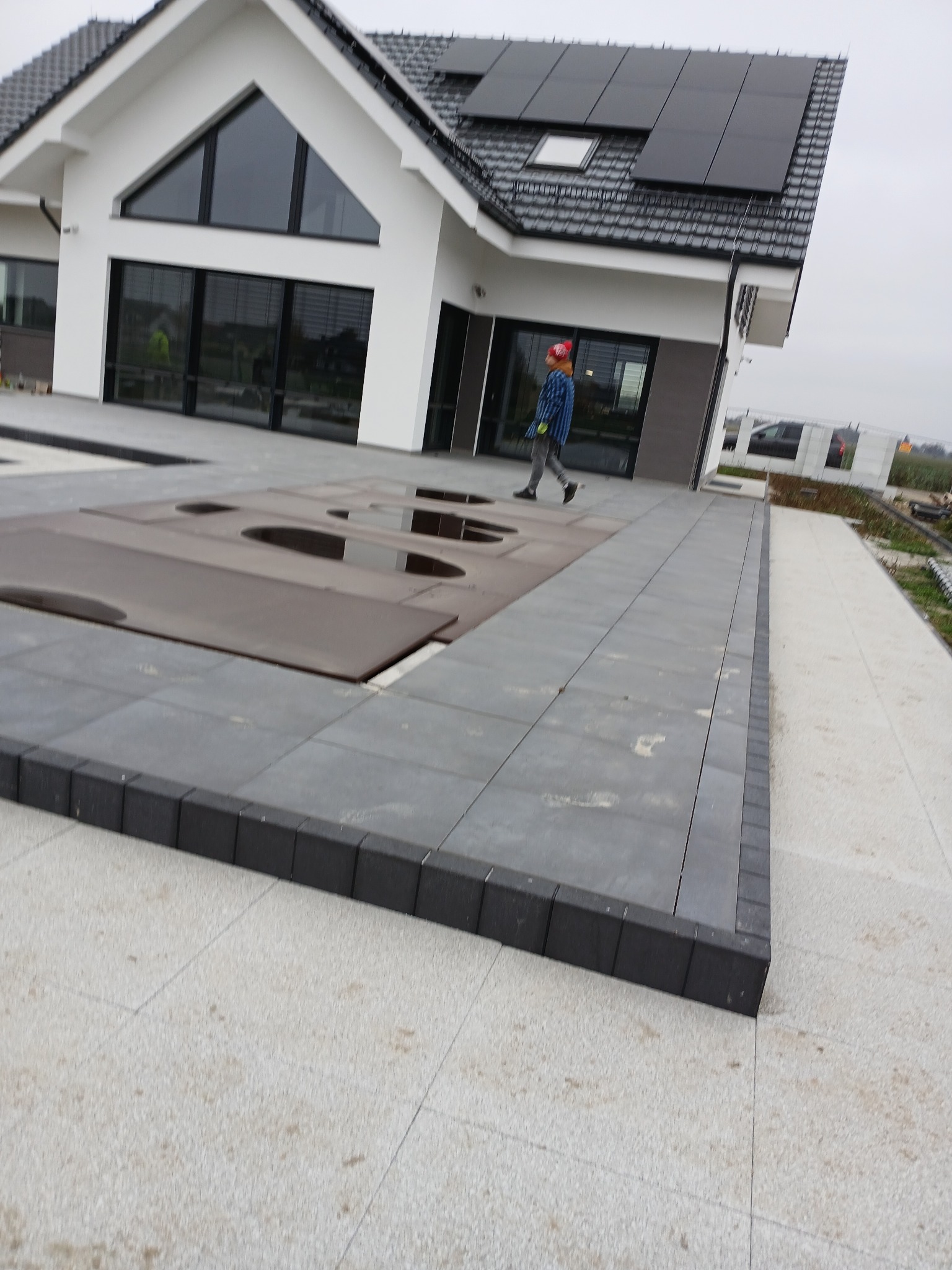 Nowoczesny dom z panelami słonecznymi i tarasem wyłożonym szarą kostką brukową, widoczny pracownik w czerwonym kasku przechodzący obok przykrytej części basenu.