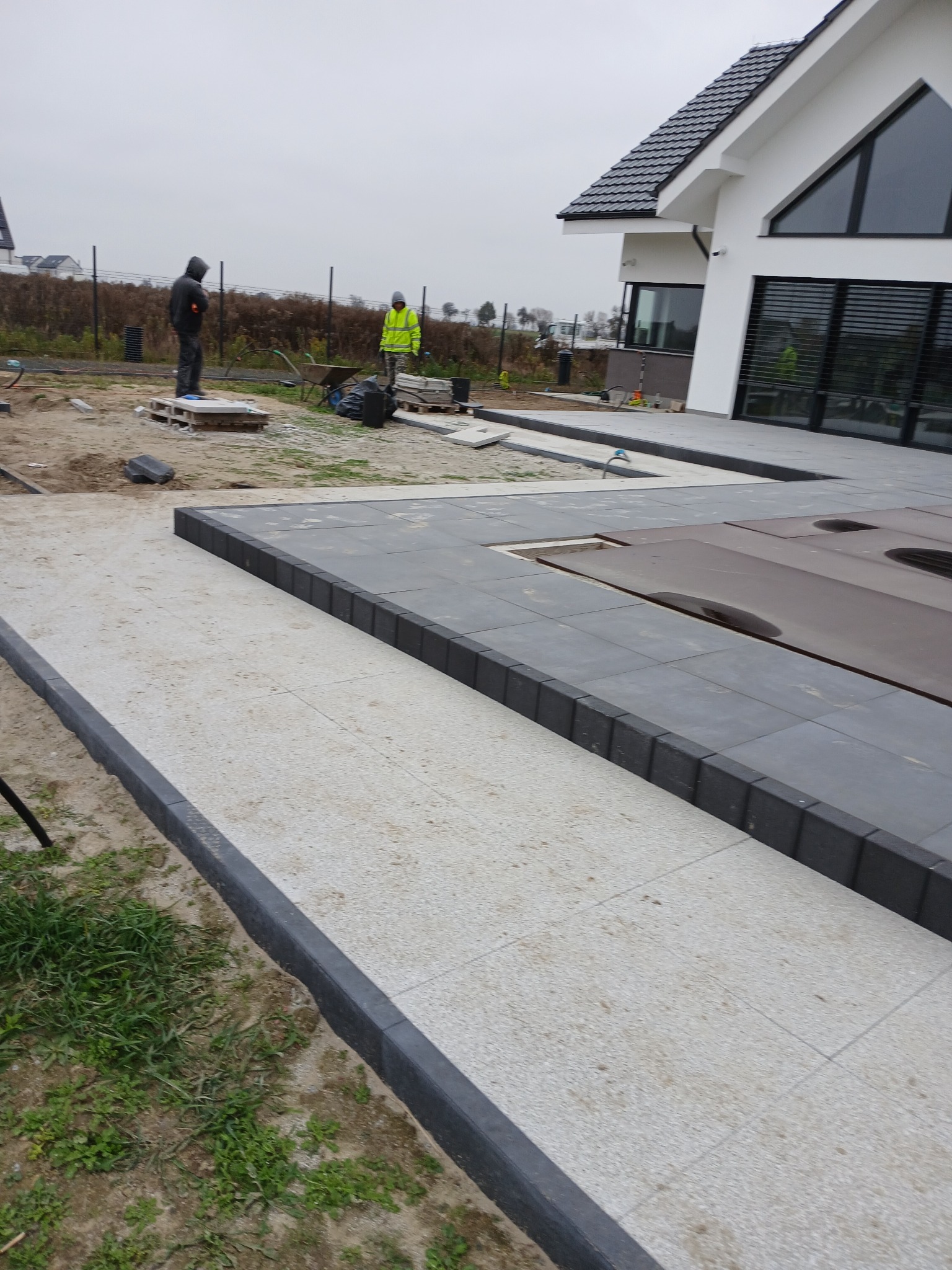 Jasnoszara nawierzchnia z obrzeżami z ciemnych kostek, w tle pracownicy i nowoczesny dom z dużymi oknami.