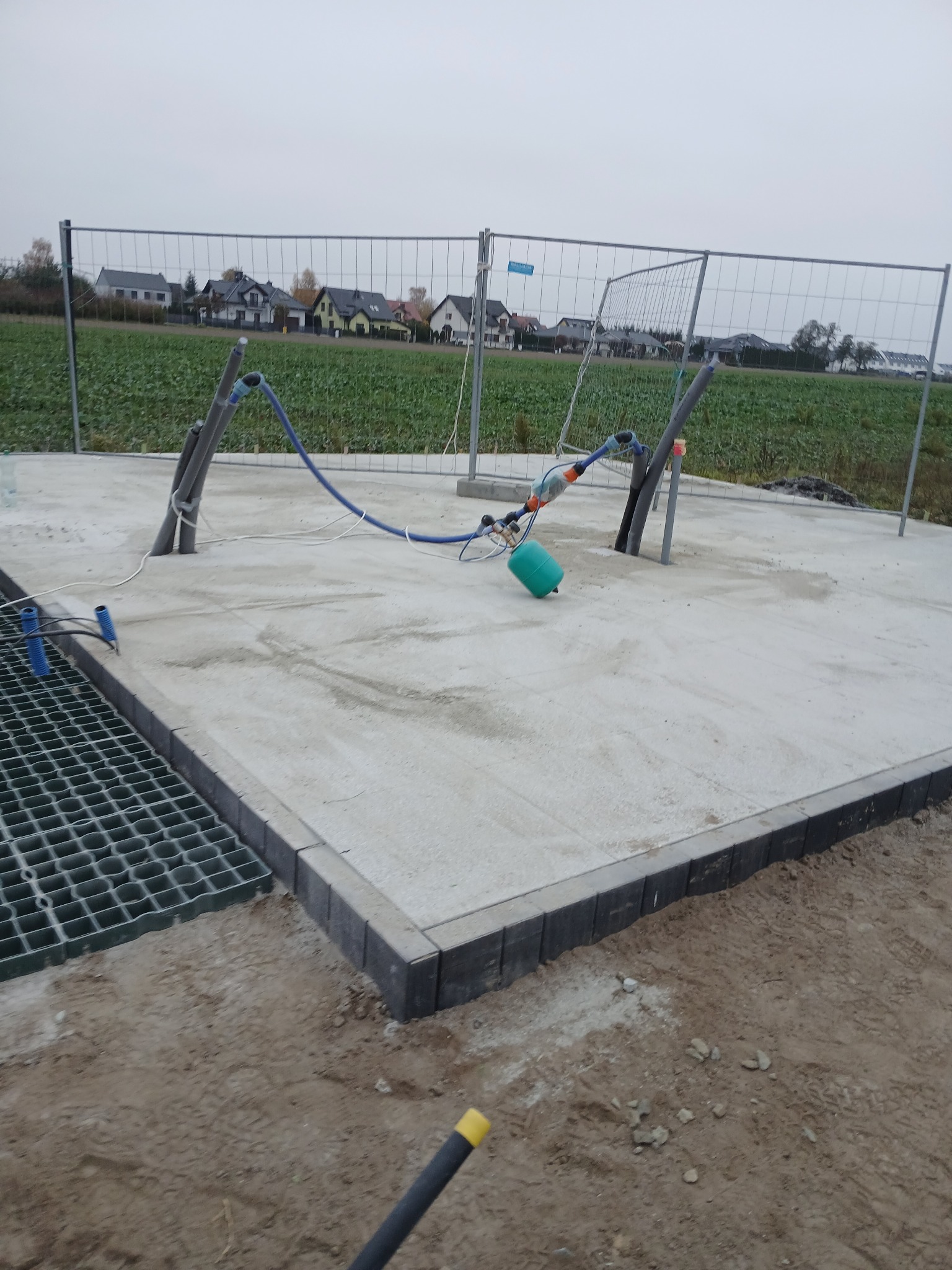 Betonowa platforma z obrzeżami z kostki, zabezpieczona siatką ogrodzeniową, z widocznymi rurami i elementami instalacji, na tle pola i zabudowań.