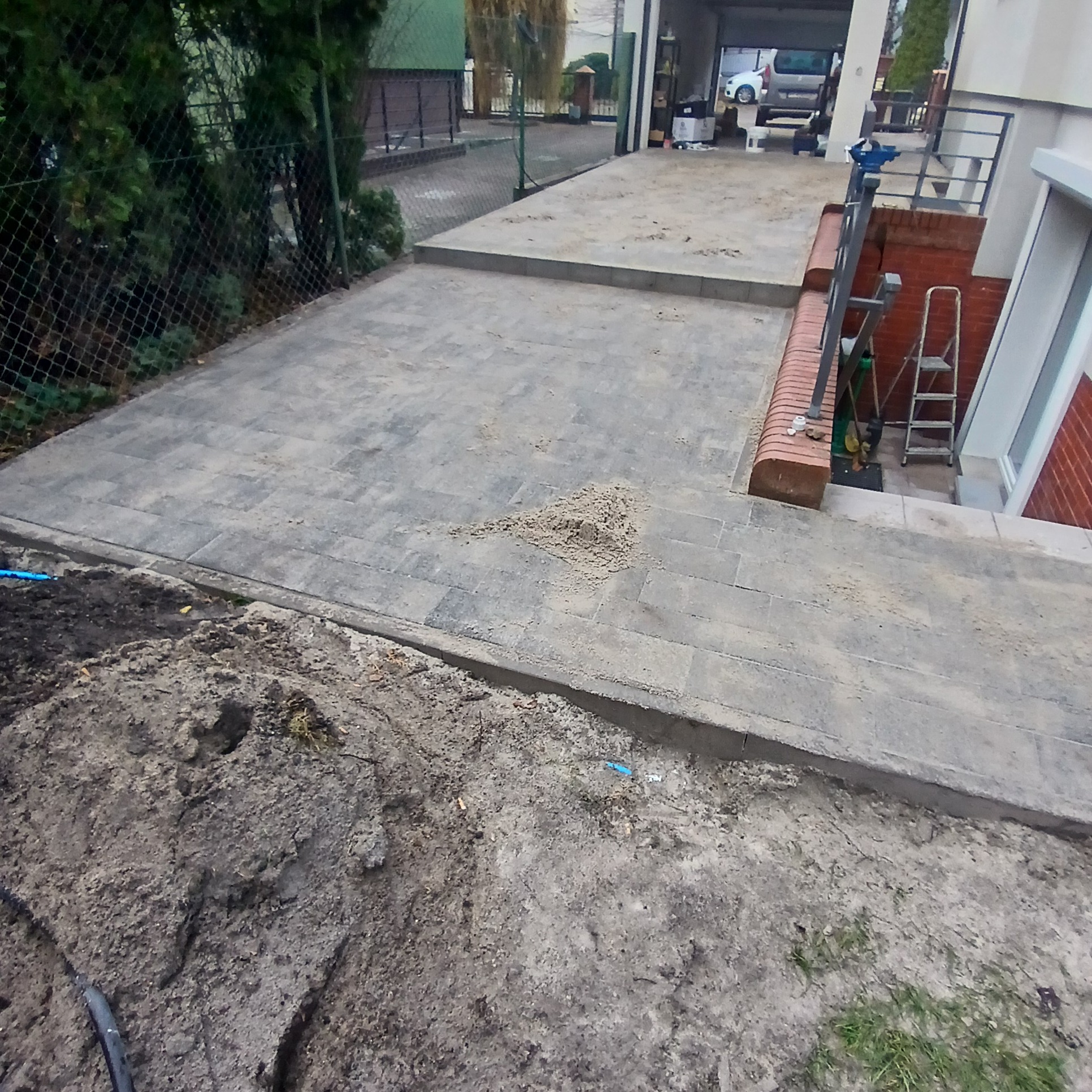 Ułożona szara kostka brukowa na podjeździe, widoczna sterta piasku, obok ziemia i trawa, w tle garaż z samochodem.