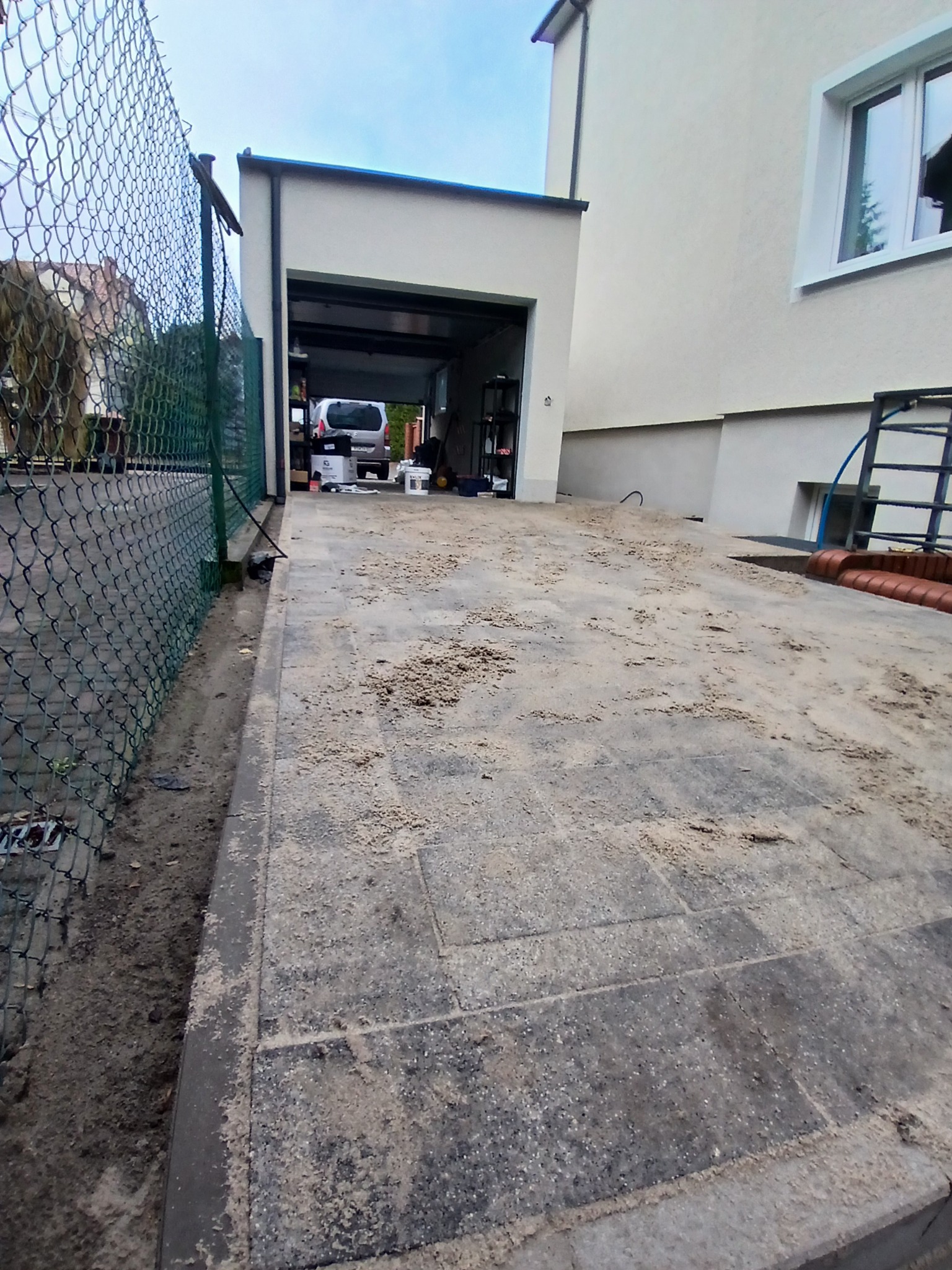 Ułożona kostka brukowa przed garażem, posypana piaskiem, widoczny fragment ogrodzenia z siatki i fragment domu z oknem.