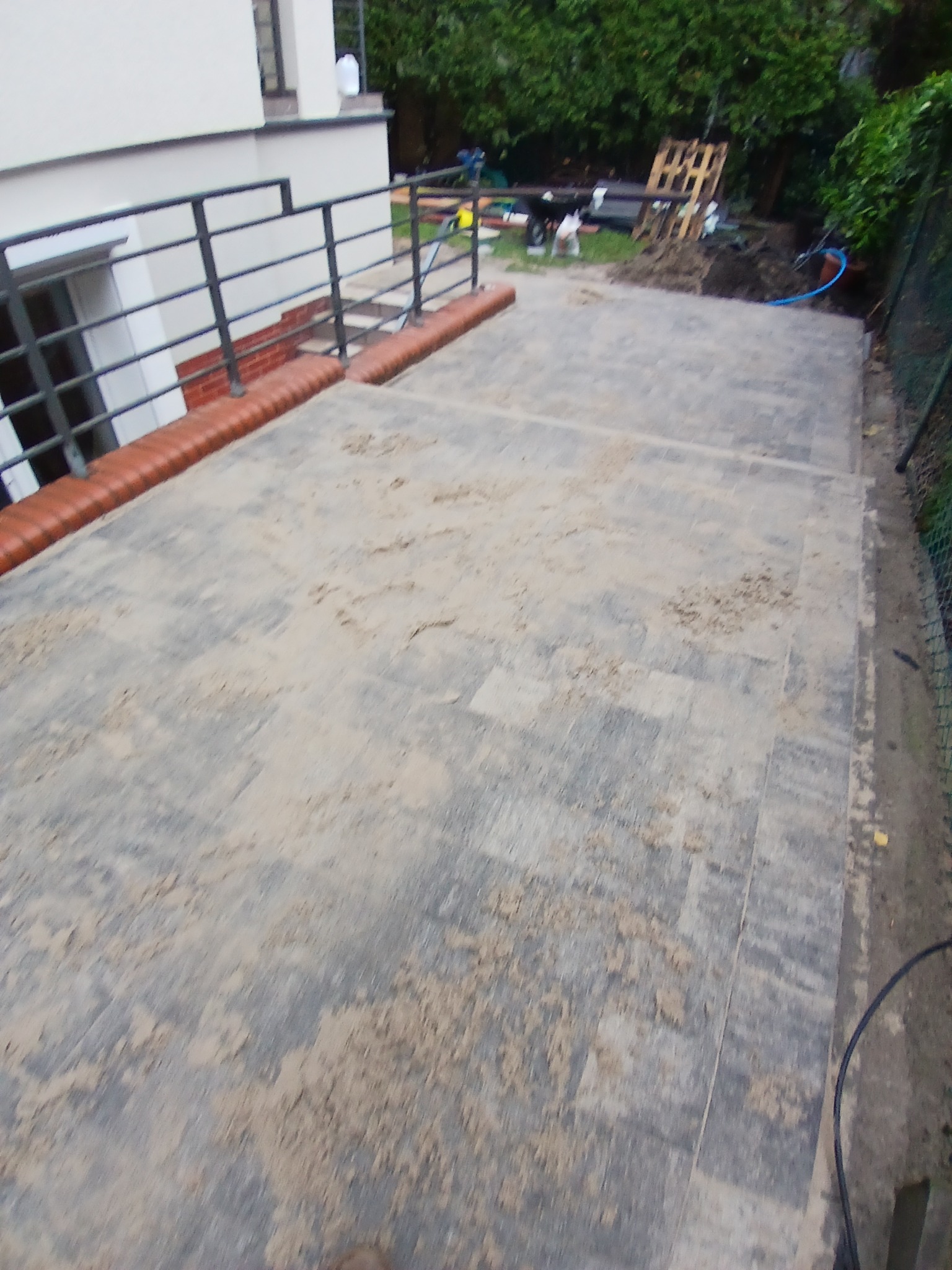 Nowo ułożona, szara kostka brukowa na tarasie z czarną balustradą i ceglanym obrzeżem, widoczne ślady piasku i zaplecze budowy w tle.
