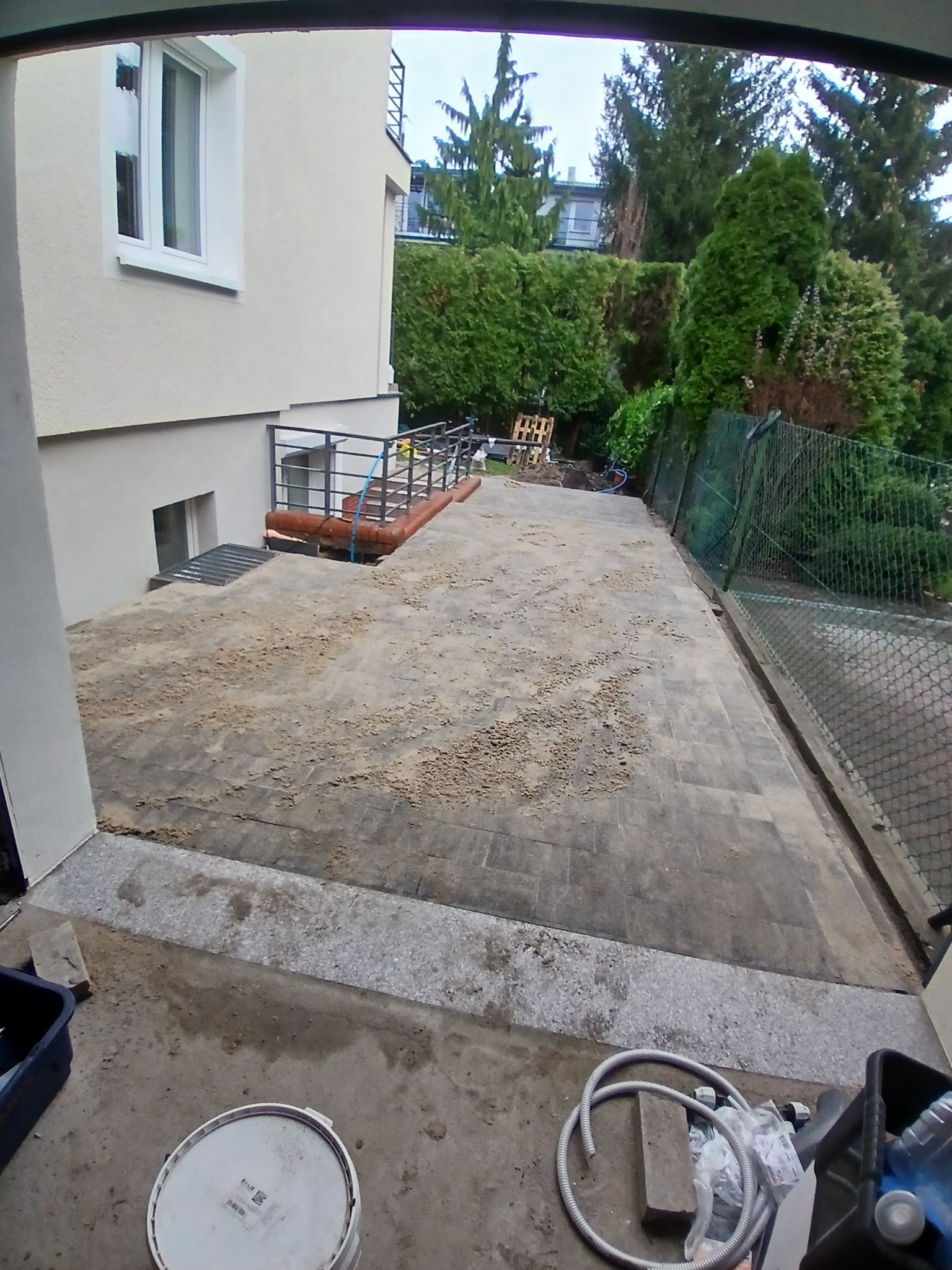 Ułożona szara kostka brukowa na posesji, częściowo zasypana piaskiem, z widoczną balustradą i elementami ogrodu w tle.