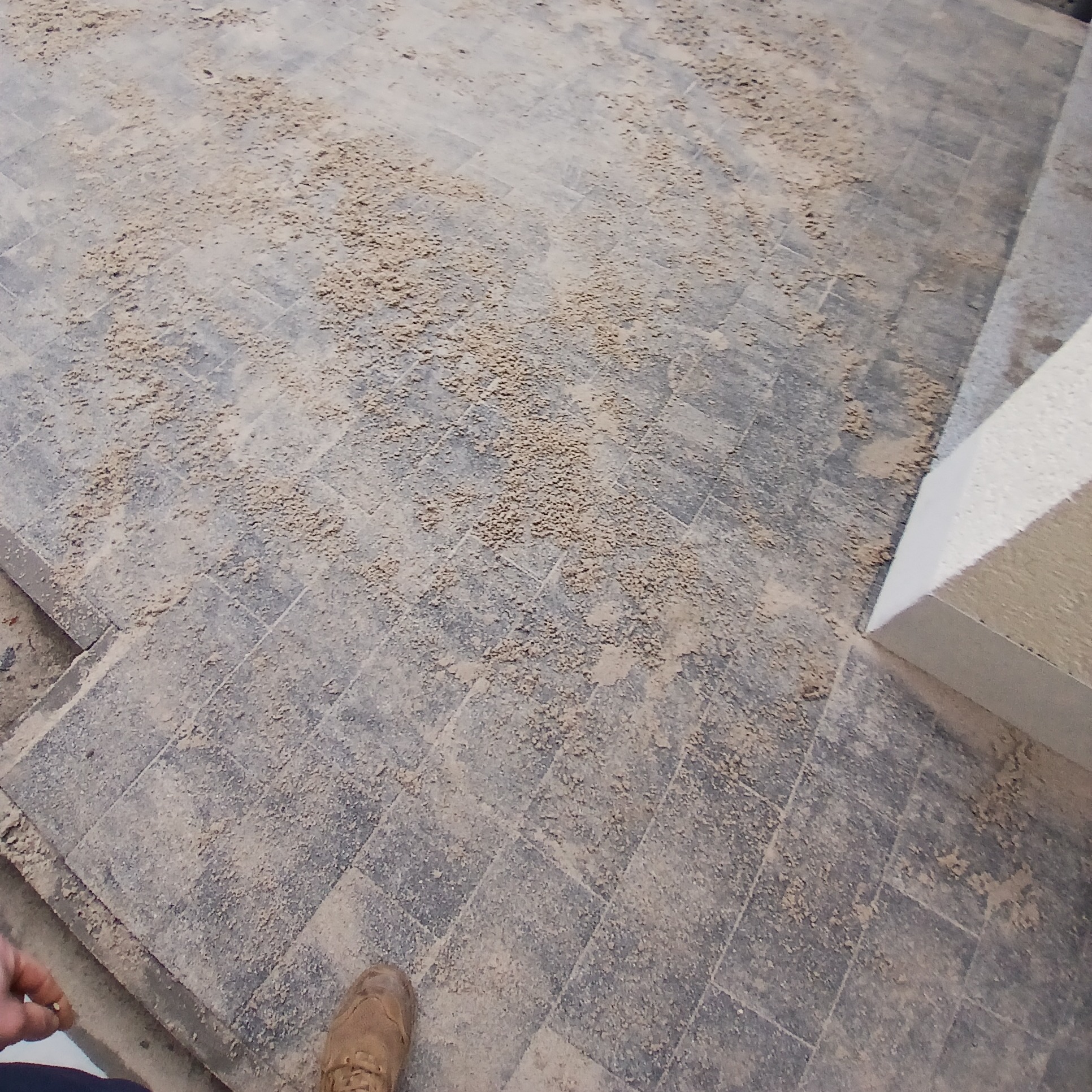 Szare kostki brukowe pokryte piaskiem, widoczny fragment buta roboczego i biały element architektoniczny, prawdopodobnie schody.