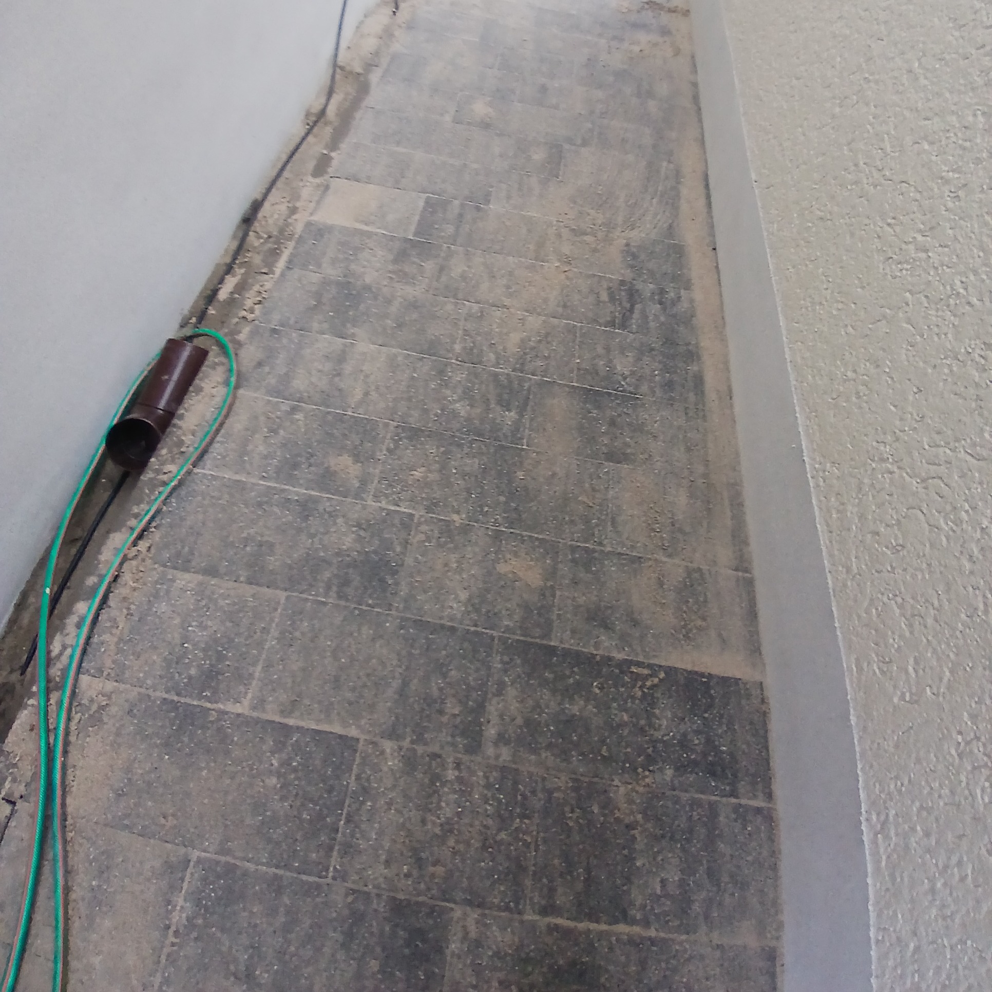 Ułożona, szara kostka brukowa przylegająca do ściany budynku. Widoczny zielony wąż i brązowa rura na powierzchni kostki.