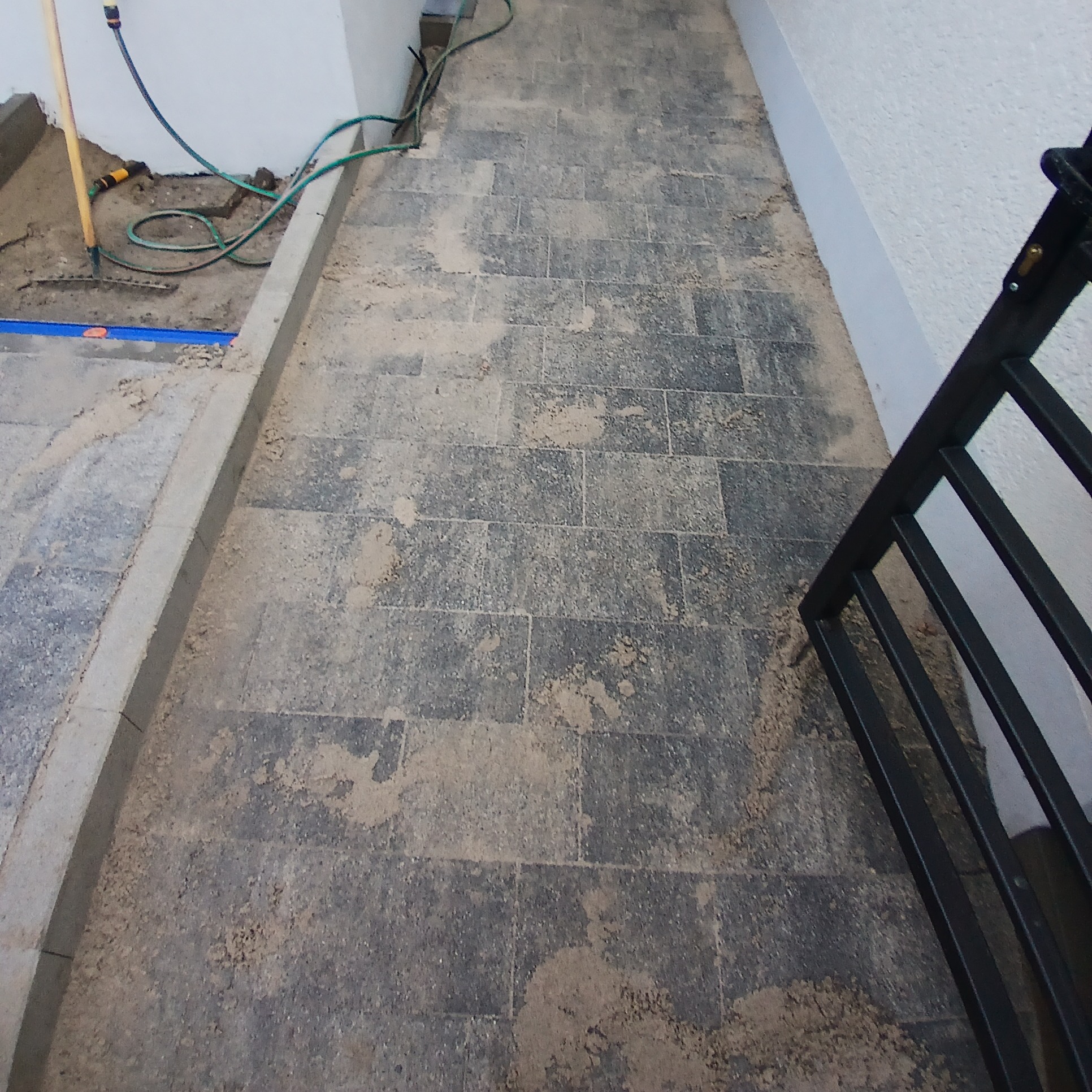 Układanie szarej kostki brukowej w trakcie prac, widoczne narzędzia: grabie i poziomica, piasek rozsypany na powierzchni, fragment czarnej barierki.